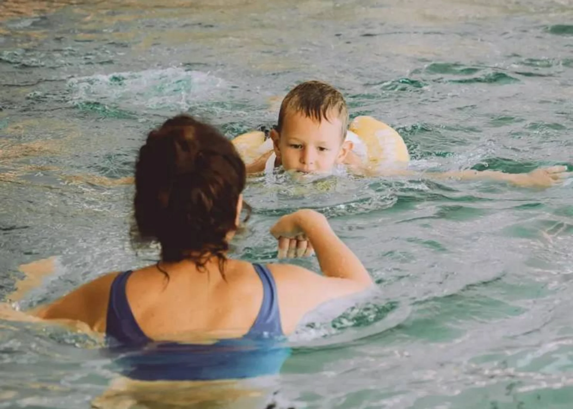 Ab ins Familienhotel mit Schwimmkurs! Frau unterrichtet Kind mit Schwimmhilfe im Hallenbad