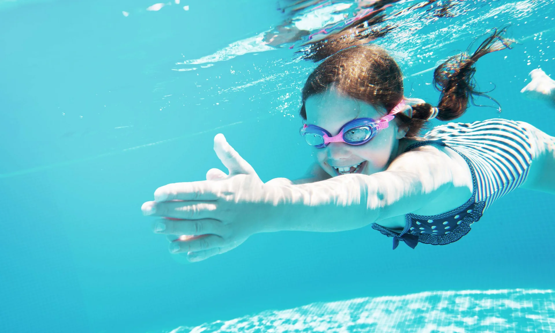 Freizeitkurse für Ihre Kids und Teenies Lächendes Mädchen schwimmt unter Wasser im Pool mit Schwimmbrille und Badeanzug