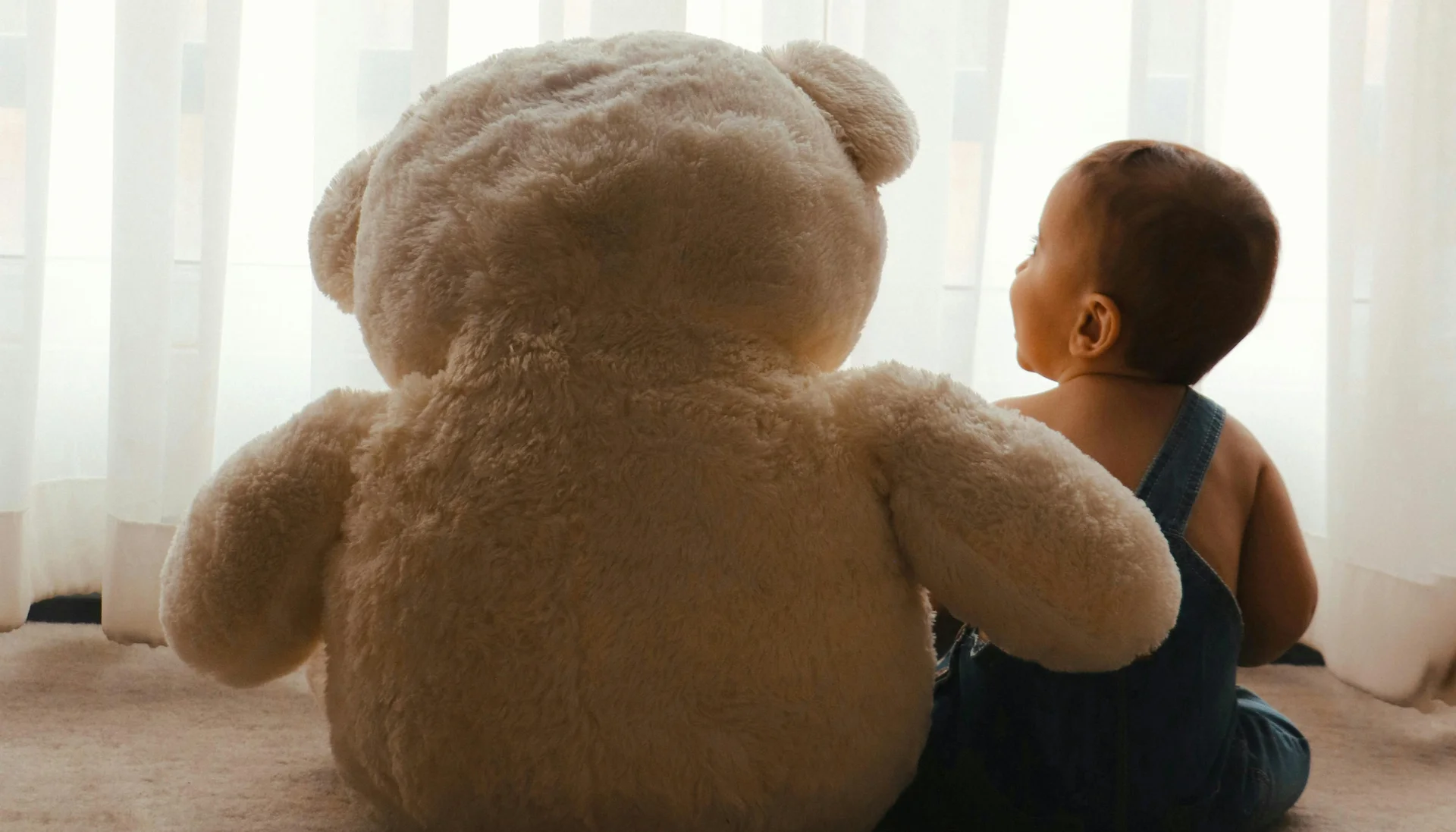 Familienurlaub mit Baby und Kleinkind im Buchau Baby sitzt neben großem Plüschbär vor weißem Vorhang