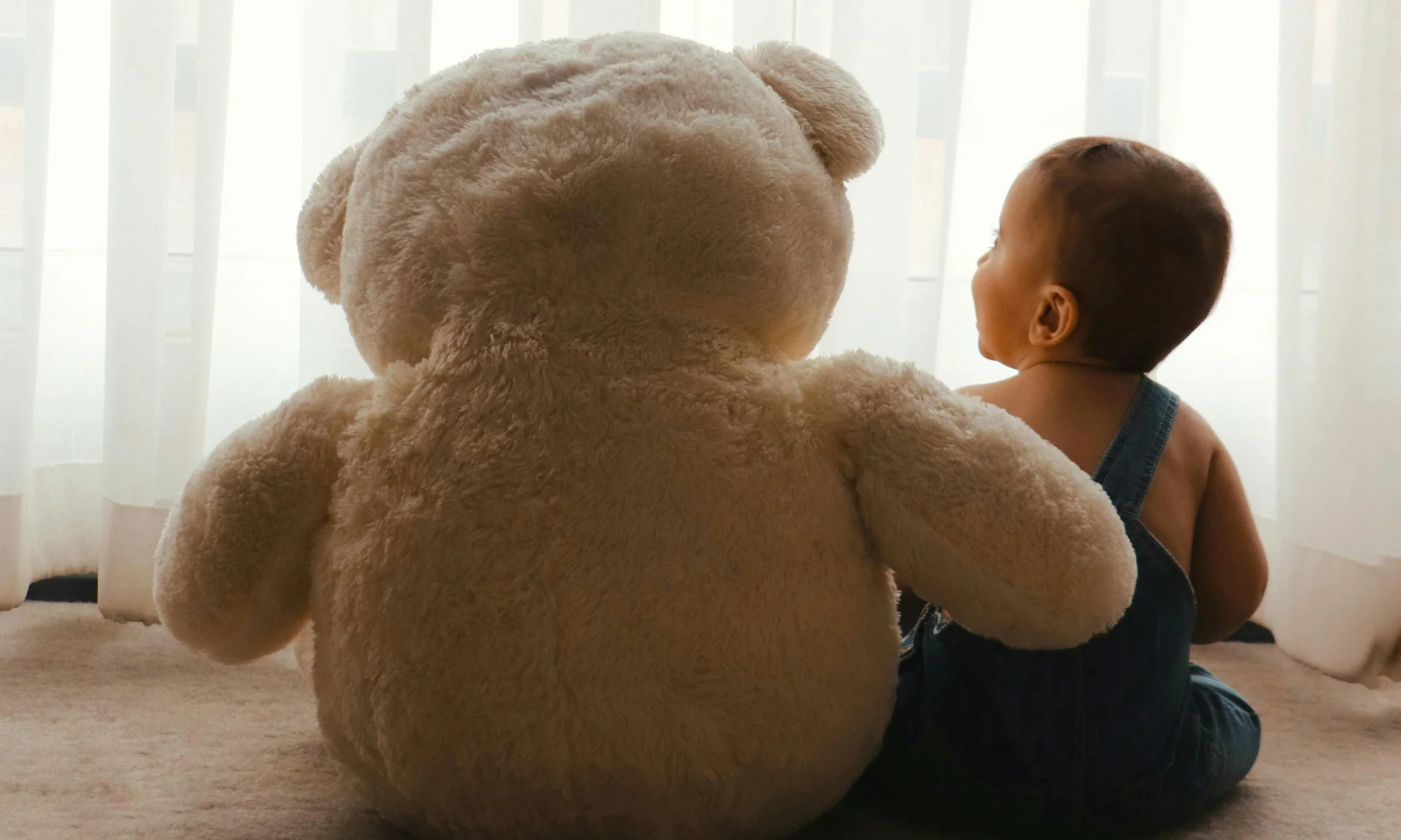 Ihr Hotel mit Kinderbetreuung in Tirol Baby sitzt neben großem Plüschbär vor weißem Vorhang