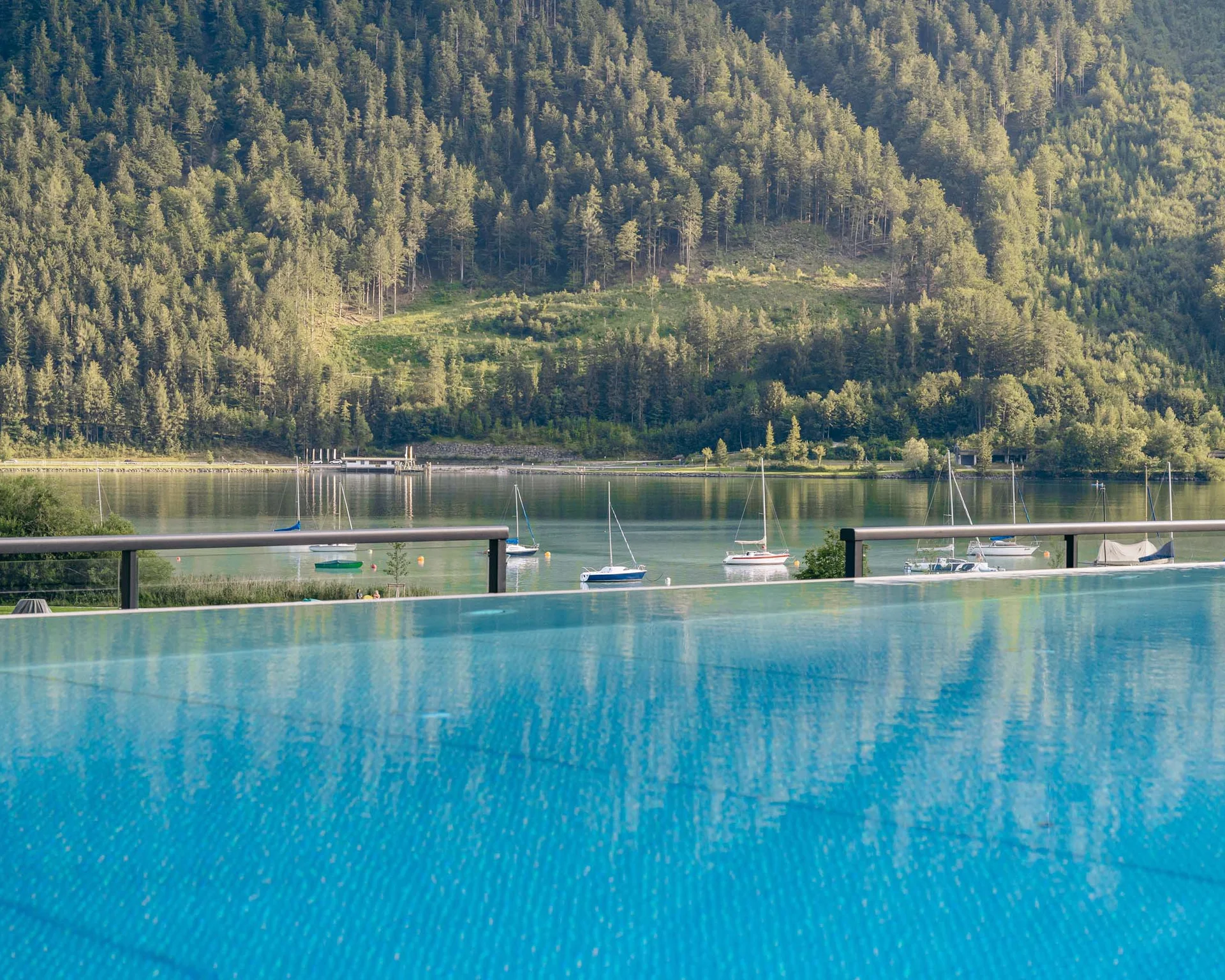 Ab ins Wellnesshotel in Tirol, in den Infinitypool! Blick auf See mit Segelbooten und bewaldeten Bergen im Hintergrund
