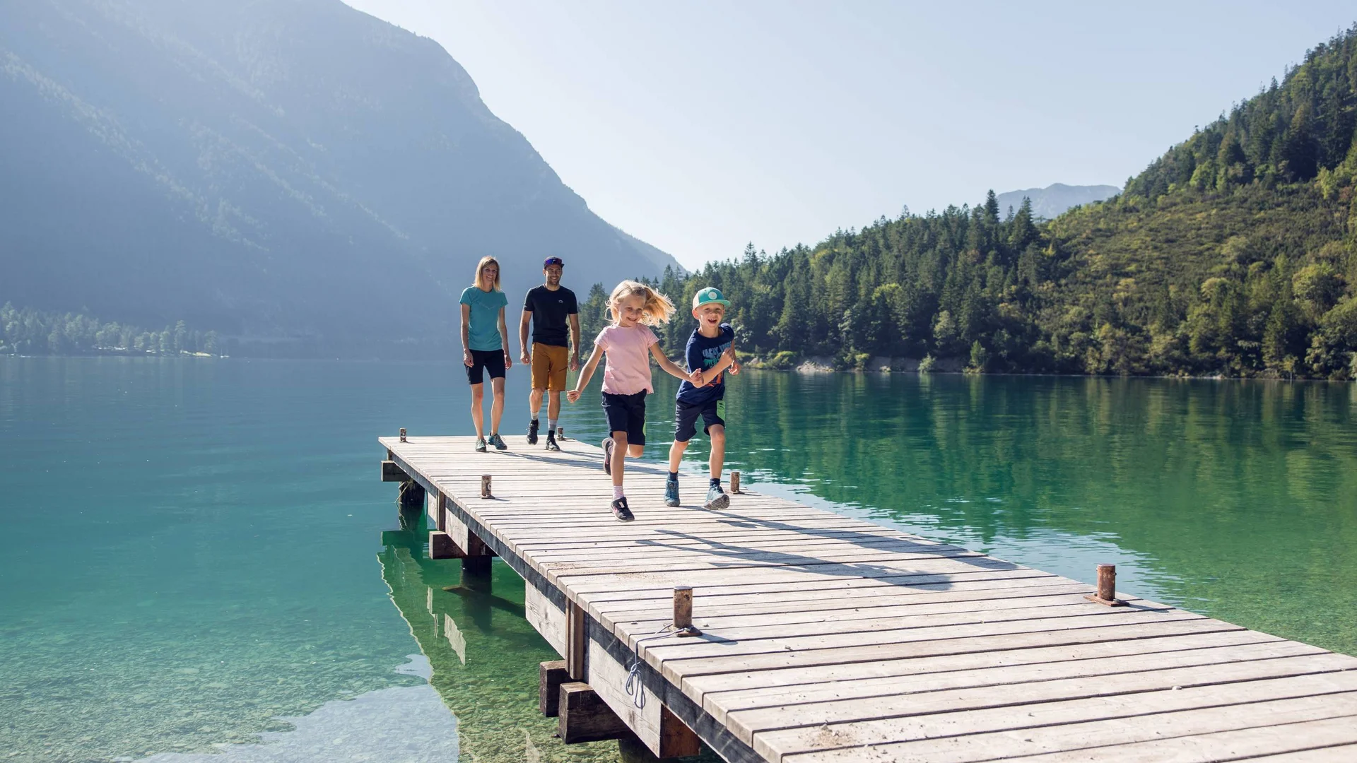 Familienzeit im Kinderhotel Buchau am Achensee Familie mit zwei Kindern läuft auf einem Steg über einem Bergsee bei Sonnenschein