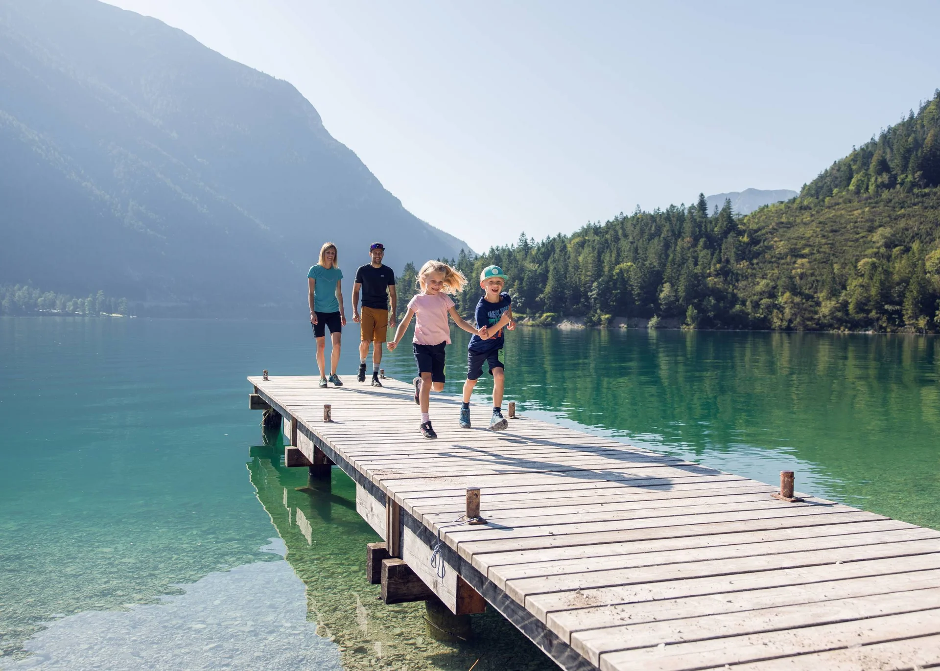Urlaub im Familienresort in Österreich Familie mit zwei Kindern läuft auf einem Steg über einem Bergsee bei Sonnenschein