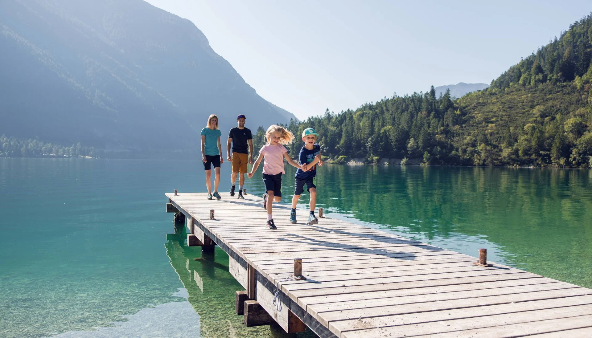 Vom Wanderhotel in Tirol direkt in die Natur Familie mit zwei Kindern läuft auf einem Steg über einem Bergsee bei Sonnenschein
