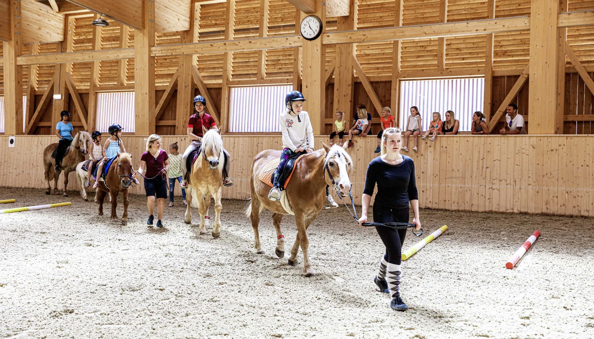 Ihr abenteuerlicher Erlebnisurlaub in Österreich Kinder beim Reitunterricht in einer hellen Holzhalle, geführt von Erwachsenen