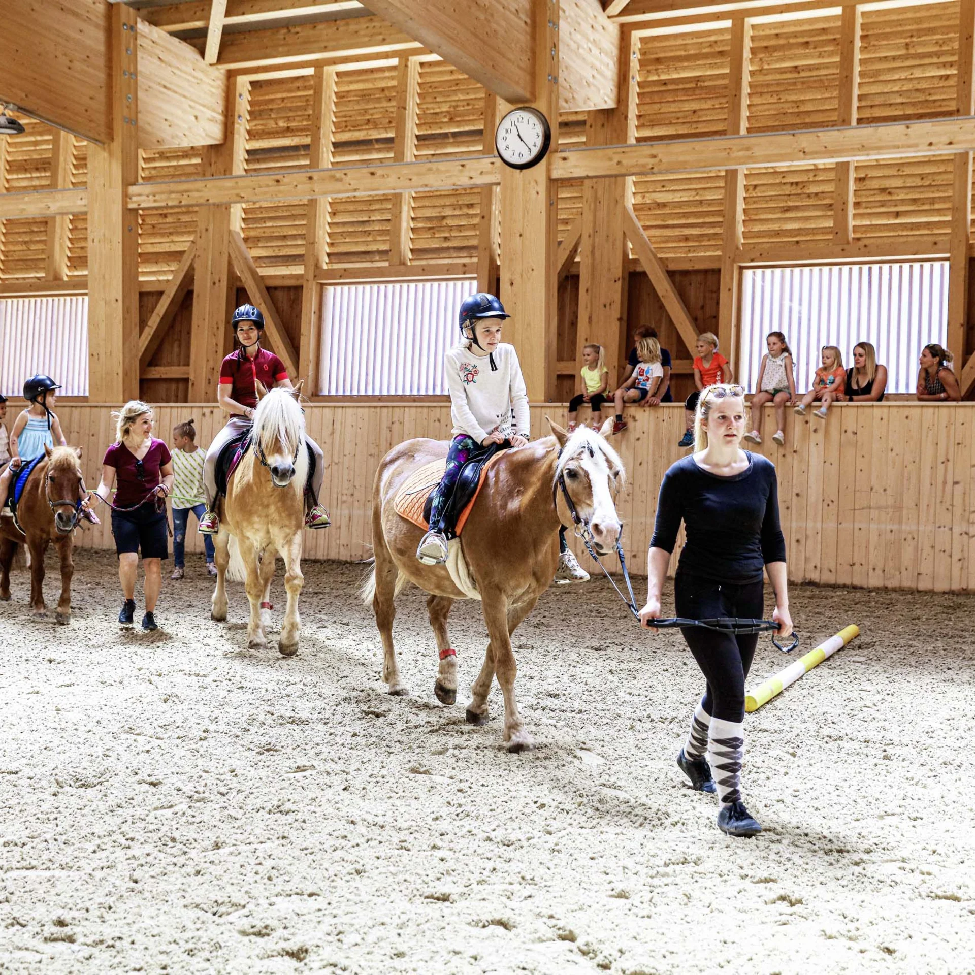 Familienzeit im Kinderhotel Buchau am Achensee Kinder beim Reitunterricht in einer hellen Holzhalle, geführt von Erwachsenen