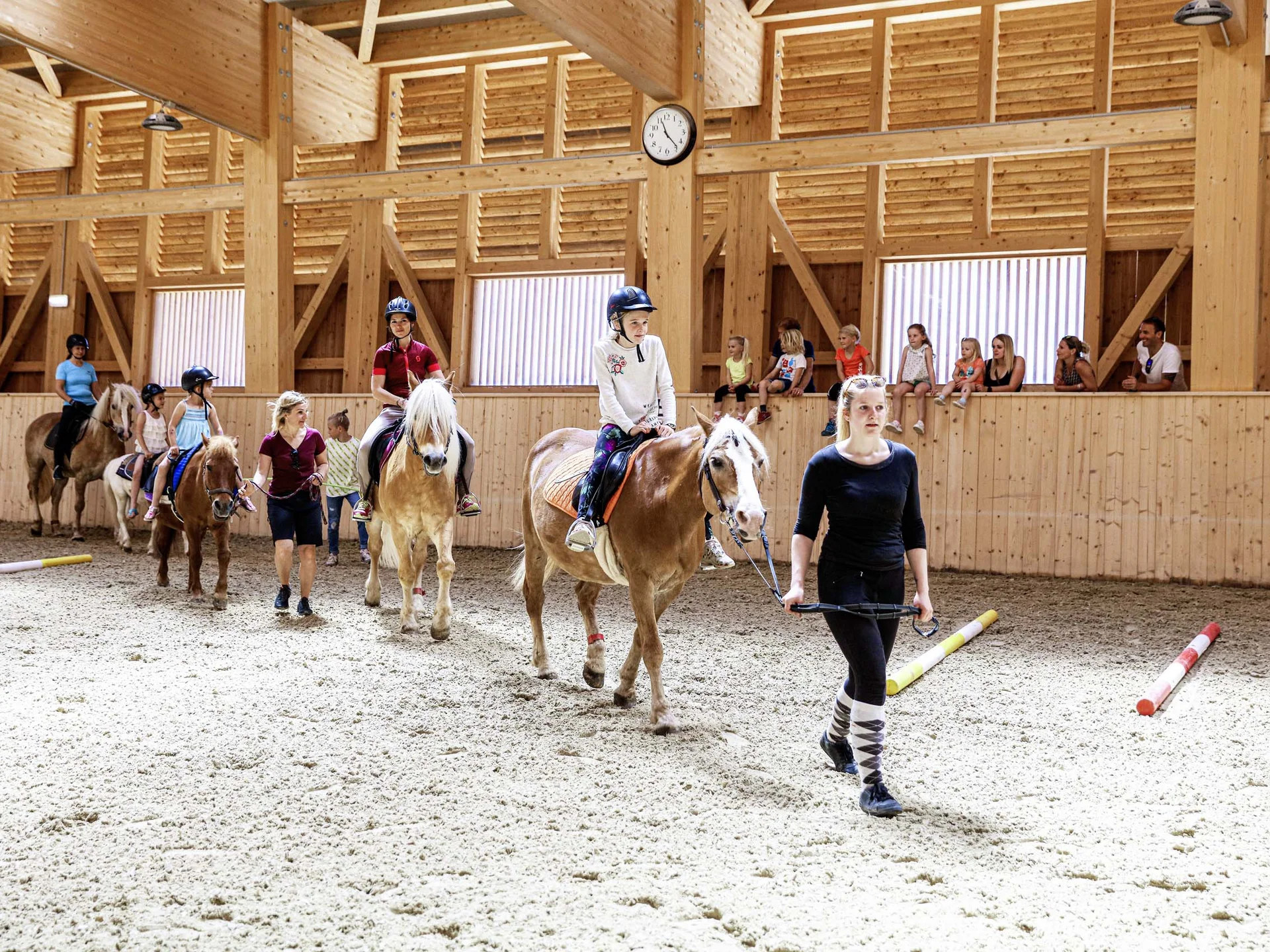Pferdeglück im Reithotel in Österreich Kinder beim Reitunterricht in einer hellen Holzhalle, geführt von Erwachsenen