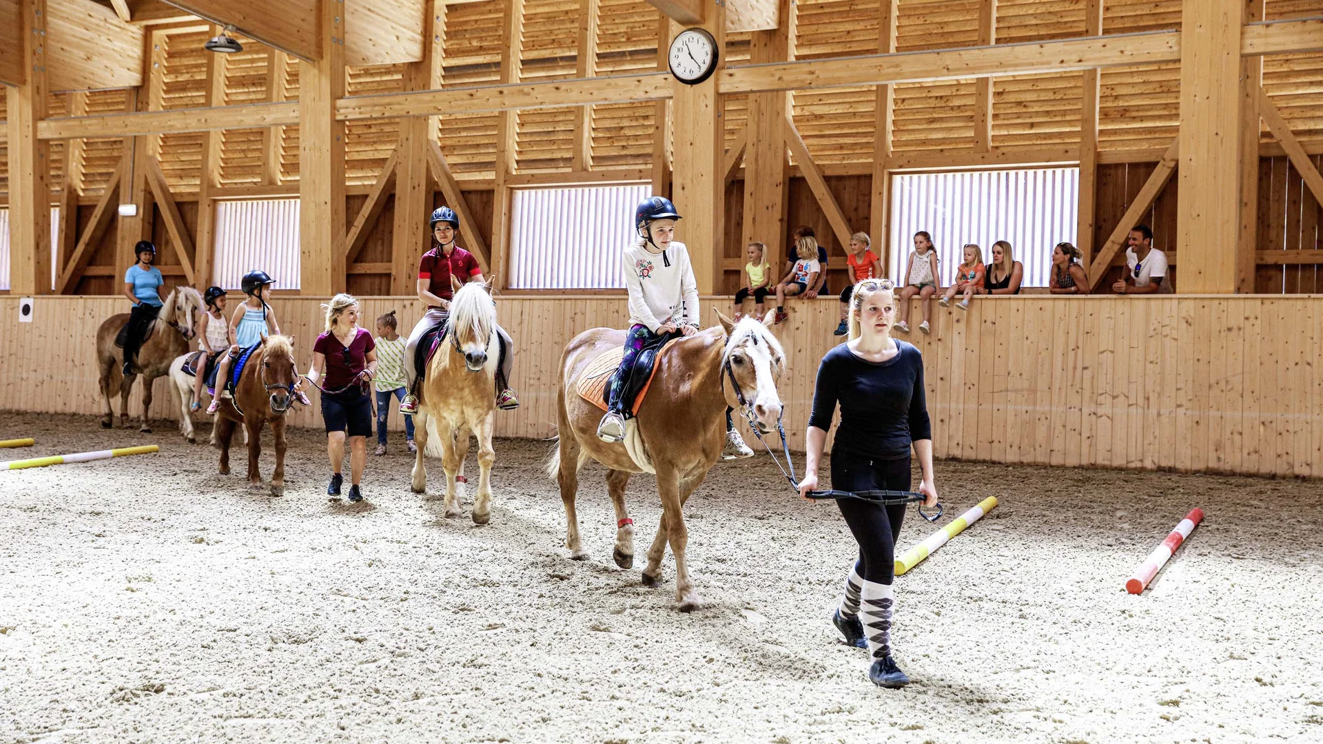 Outdoorspaß im Familienhotel am See in Österreich Kinder beim Reitunterricht in einer hellen Holzhalle, geführt von Erwachsenen
