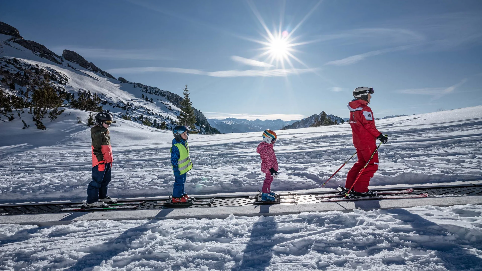 Freizeitkurse für Ihre Kids und Teenies Skilehrer mit drei Kindern auf einem Förderband im verschneiten Skigebiet bei Sonnenschein