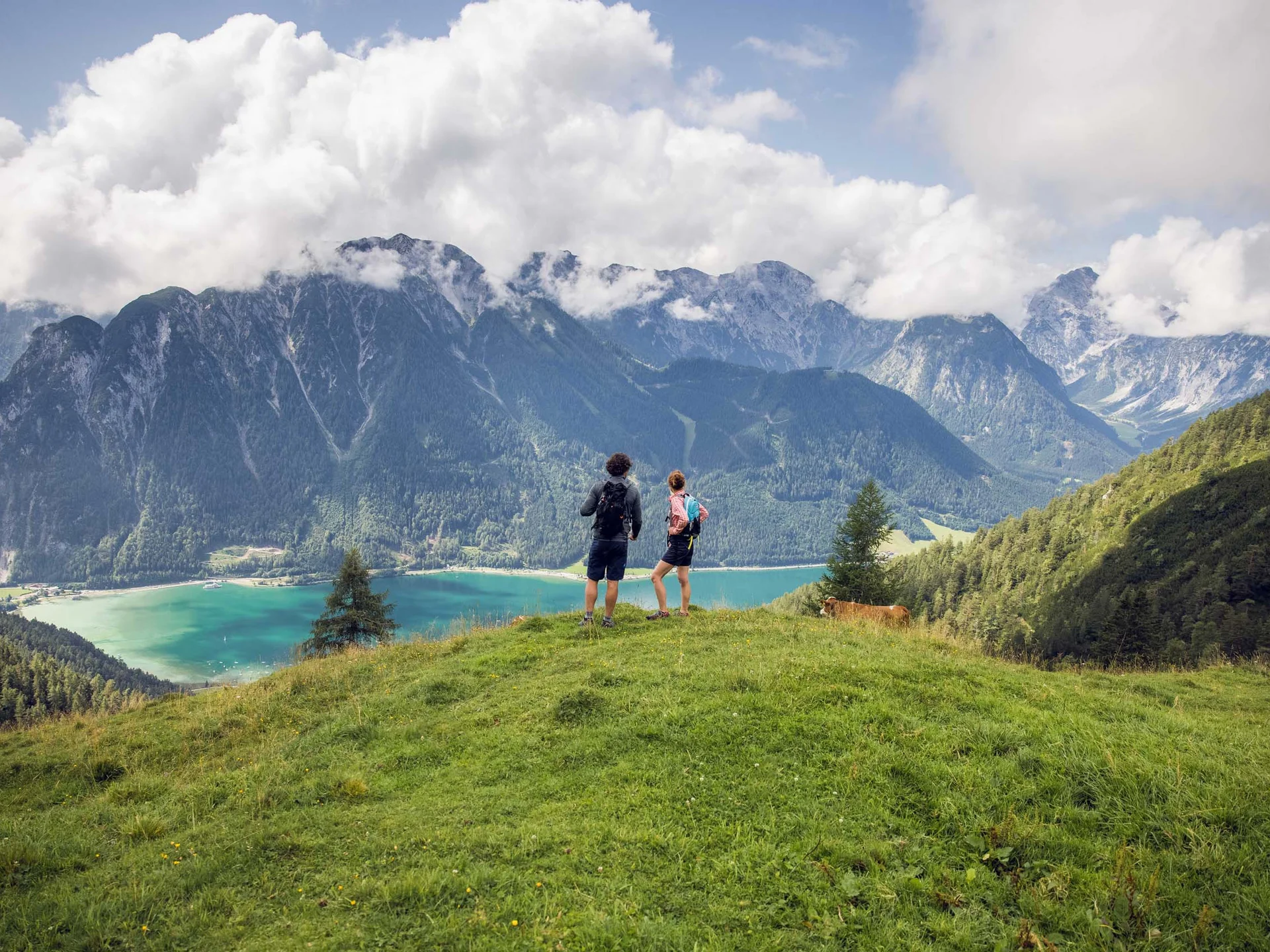Vom Wanderhotel in Tirol direkt in die Natur Zwei Wanderer stehen auf einer Wiese mit Berg- und Seeblick