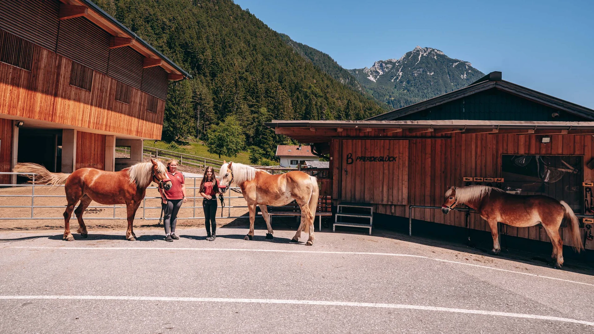 Pferdeglück im Reithotel in Österreich Zwei Mädchen mit Pferden vor Holzhütten in den Bergen bei klarem Himmel