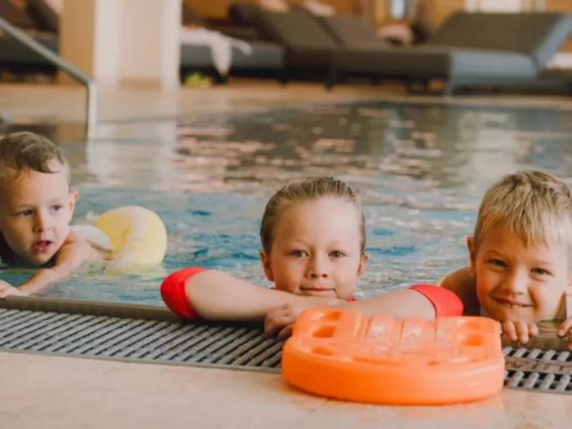 Ab ins Familienhotel mit Schwimmkurs! Drei Kinder im Hallenbad am Beckenrand mit Schwimmhilfen