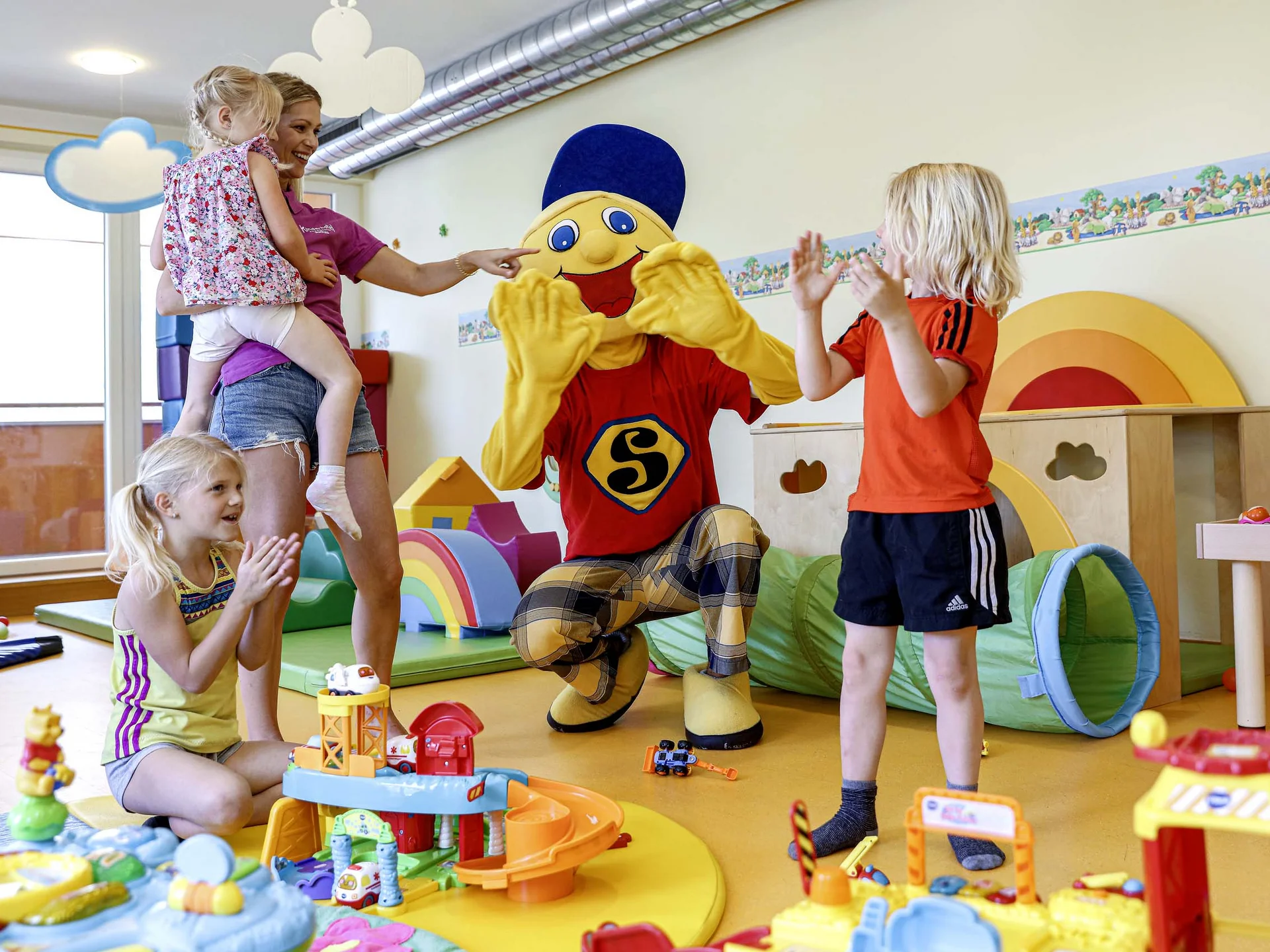 Ihr Hotel mit Kinderbetreuung in Tirol Kinder spielen und klatschen mit einer Person in einem bunten Maskottchenkostüm in einem Spielzimmer