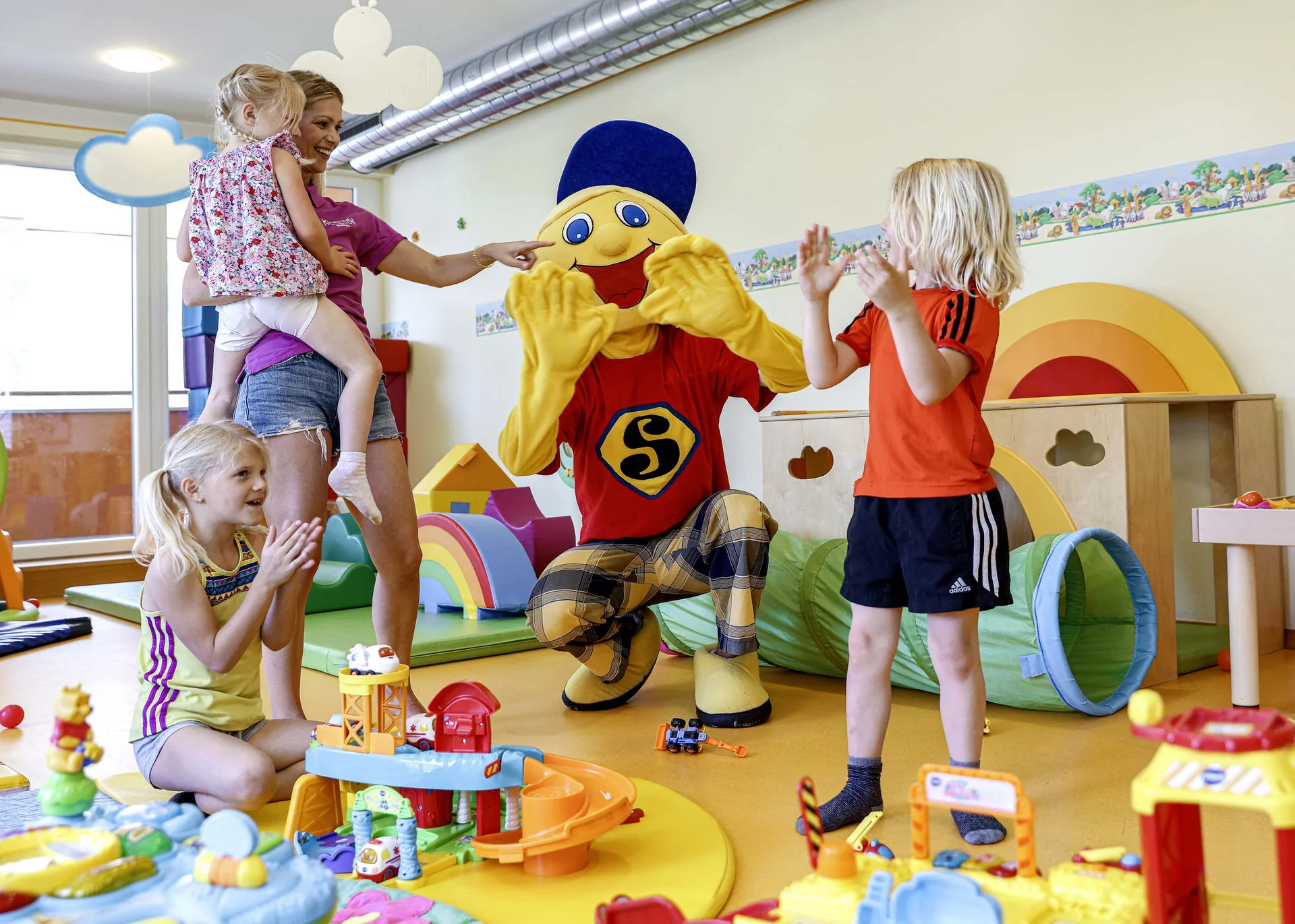 Familienzeit im Kinderhotel Buchau am Achensee Kinder spielen und klatschen mit einer Person in einem bunten Maskottchenkostüm in einem Spielzimmer