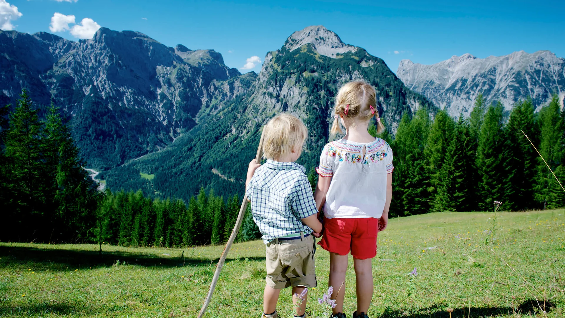 Vom Wanderhotel in Tirol direkt in die Natur Kinder in den Alpen blicken auf Berge und Wälder unter blauem Himmel