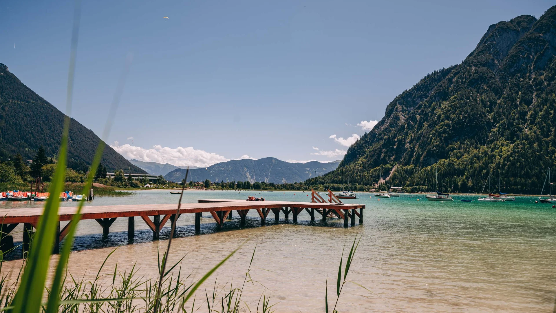 Outdoorspaß im Familienhotel am See in Österreich Holzsteg am klaren Bergsee mit Segelbooten und bewaldeten Bergen im Hintergrund