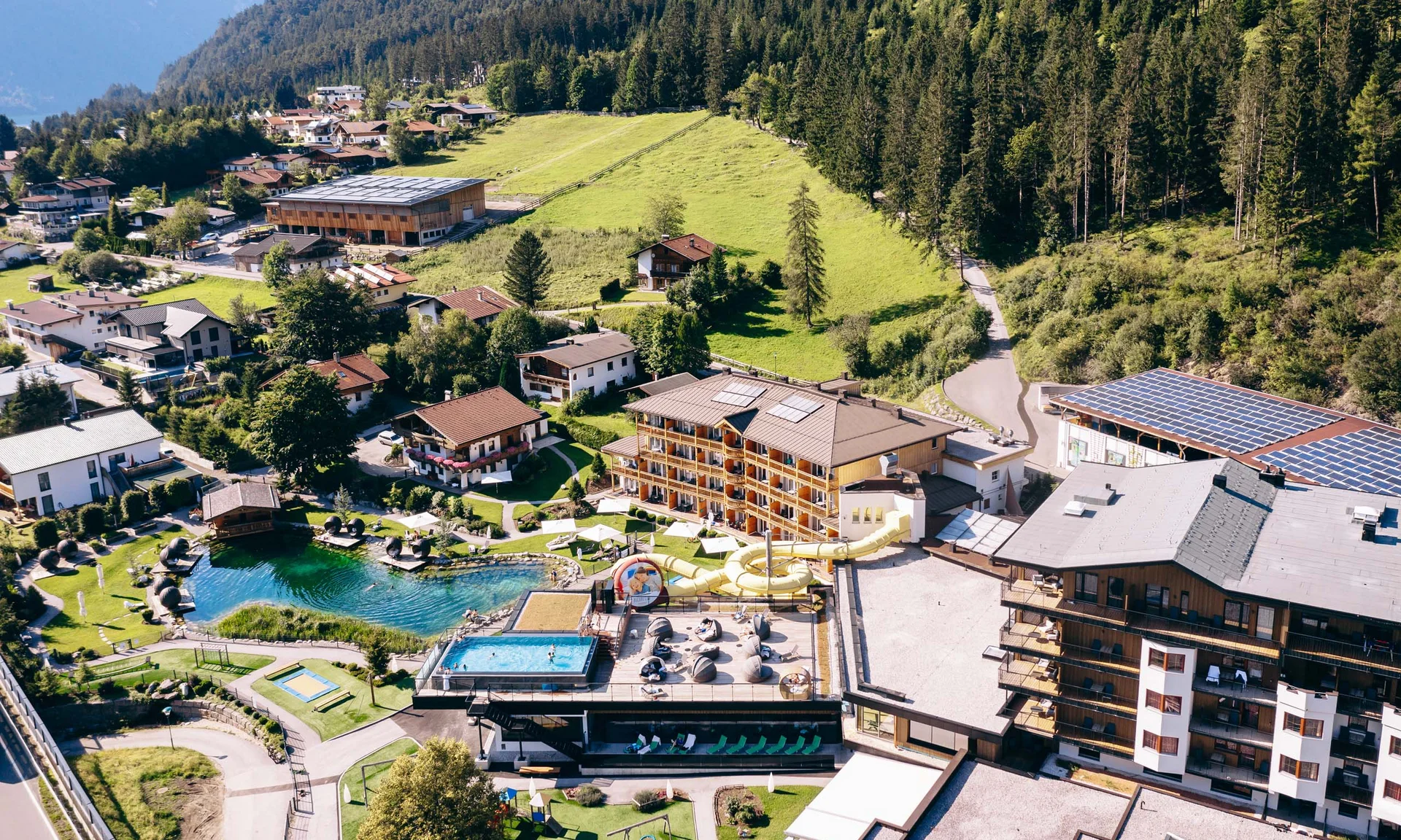 Wir lassen Bilder sprechen. Luftaufnahme eines Resorts mit Swimmingpools und Wasserrutsche in grüner Alpenlandschaft