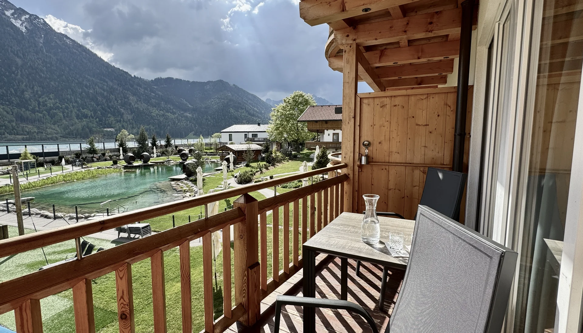Entspannte Ferien für die ganze Familie. Balkon mit Stühlen und Tisch mit Berg- und Teichblick bei bewölktem Himmel