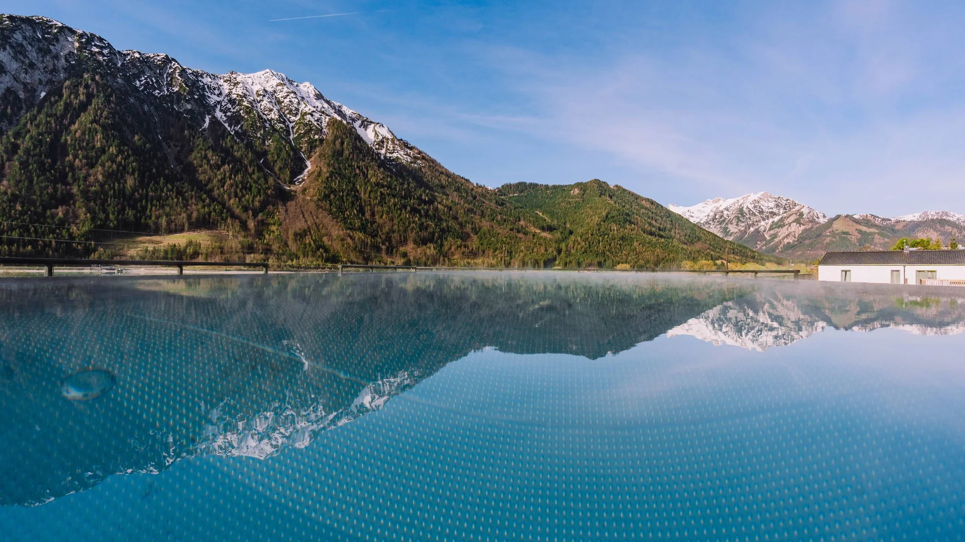 Ab ins Wellnesshotel in Tirol, in den Infinitypool! Berglandschaft mit schneebedeckten Gipfeln spiegelt sich in einem ruhigen Pool