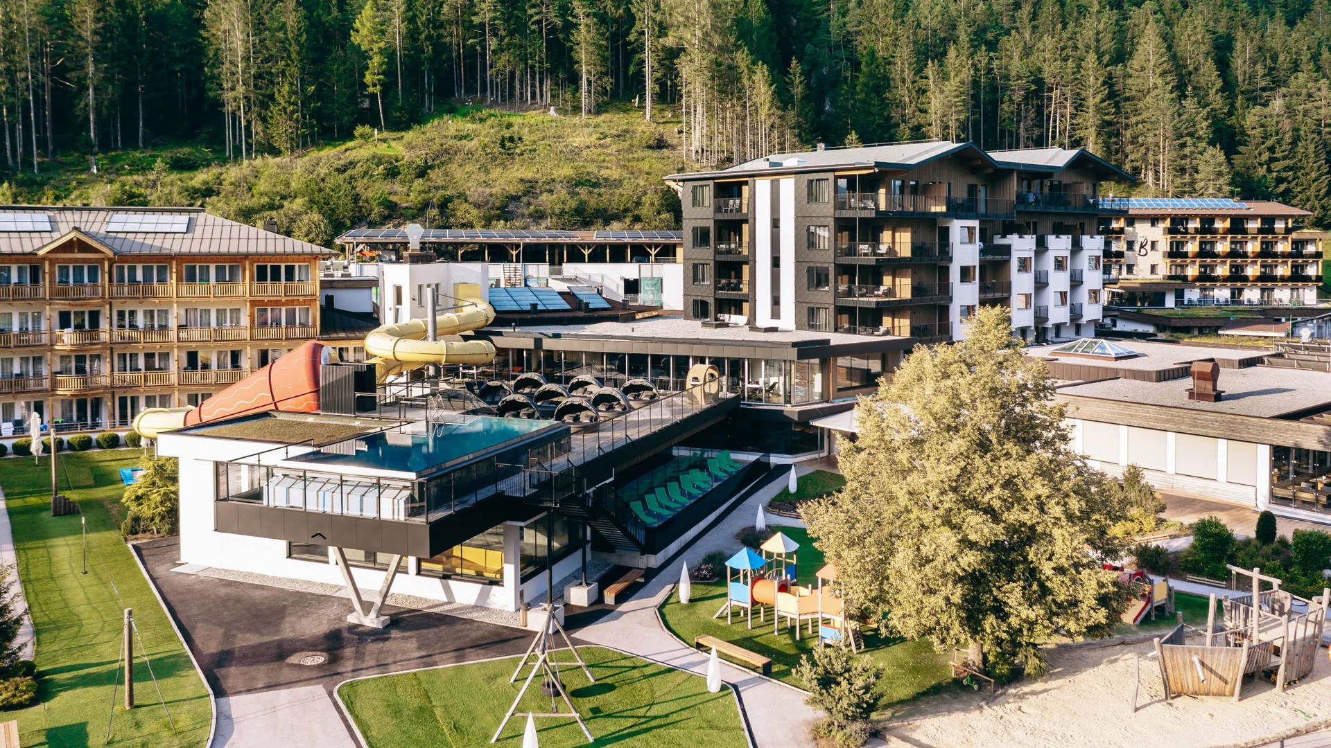 Wasserspaß im Familienhotel in Tirol mit Pool Moderner Hotelkomplex mit Wasserrutschen, Spielplatz und Wald im Hintergrund