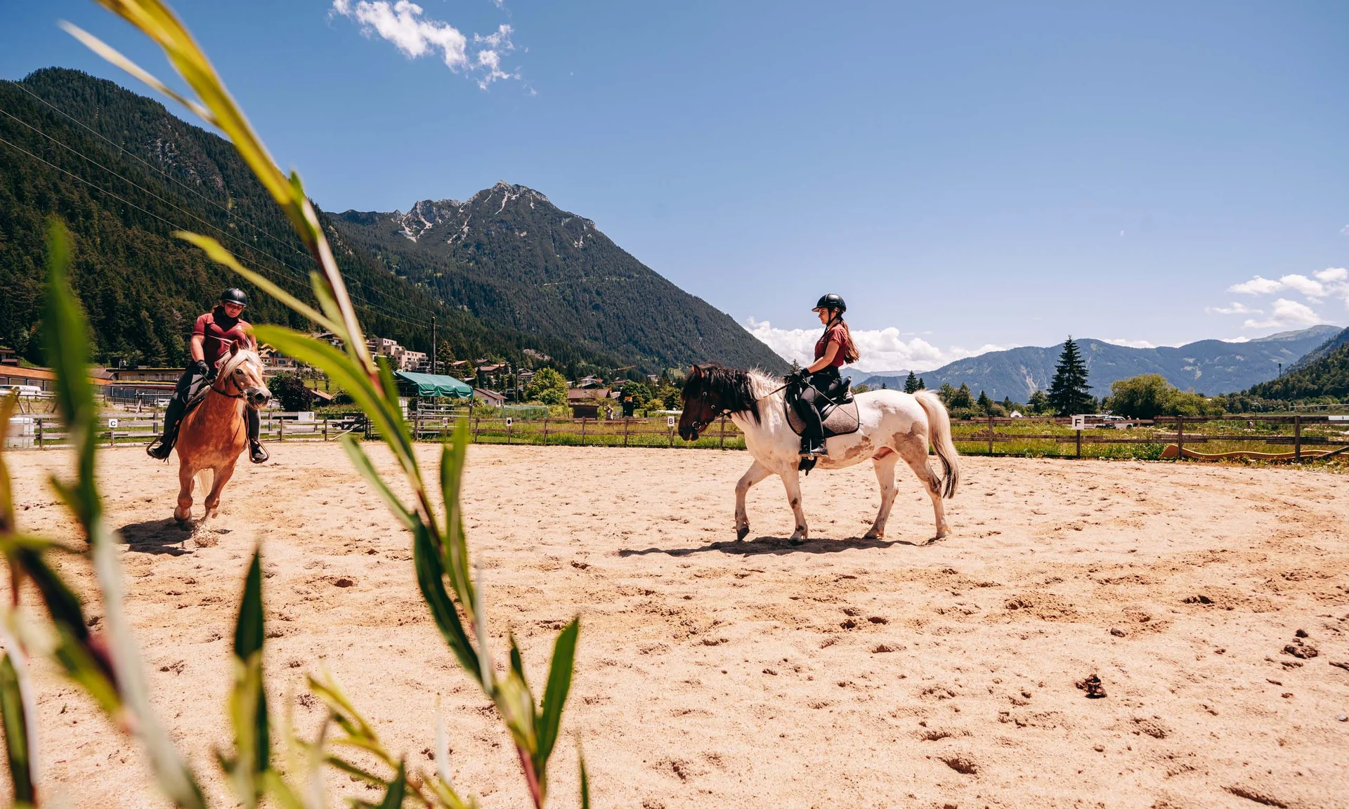 Familienurlaub mit Baby und Kleinkind im Buchau Zwei Personen reiten Pferde in einem Sandpaddock vor Bergkulisse bei Sonnenschein