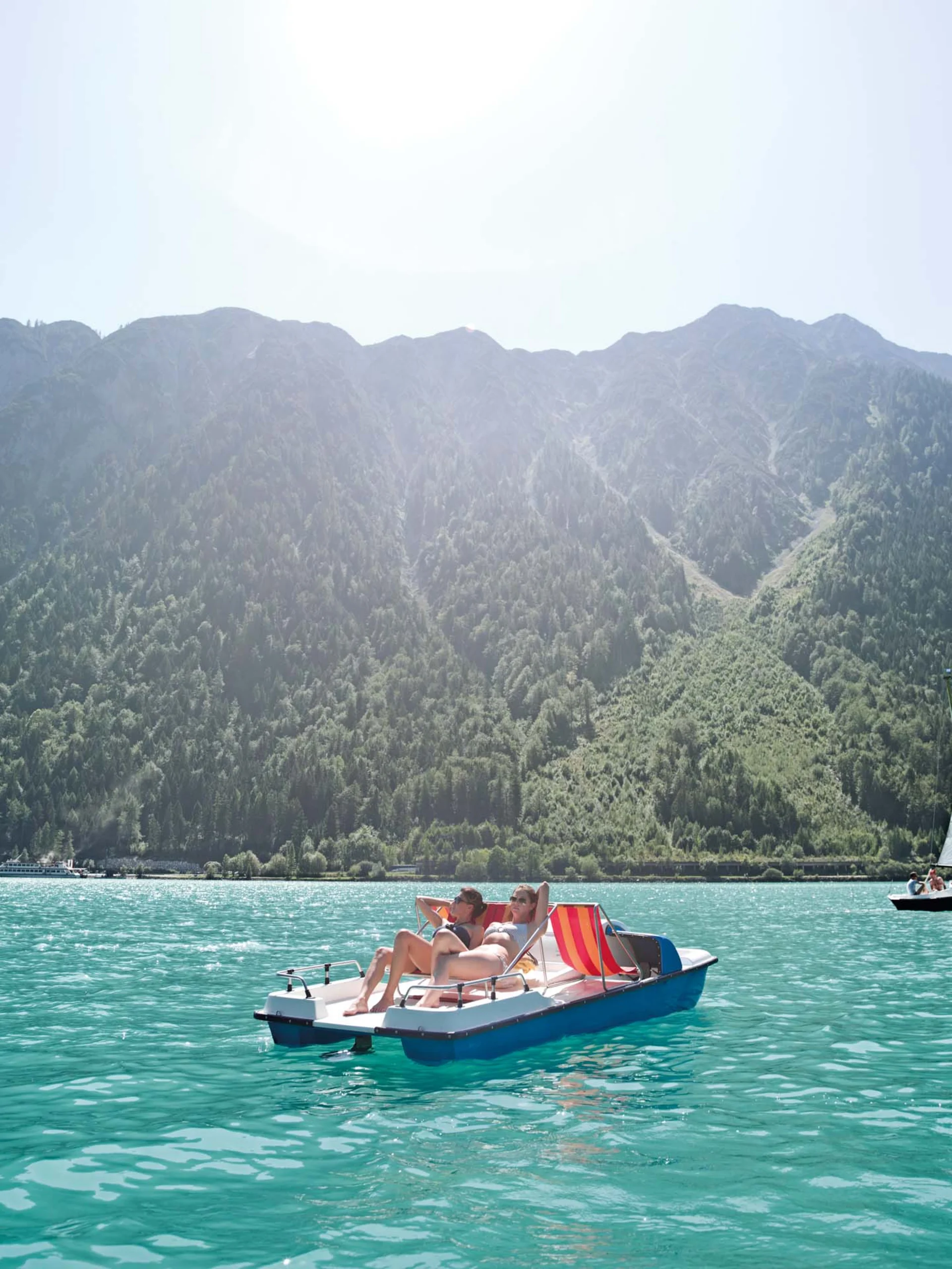 Wir lassen Bilder sprechen. Zwei Personen entspannen auf einem Tretboot auf dem türkisfarbenen See vor Bergen