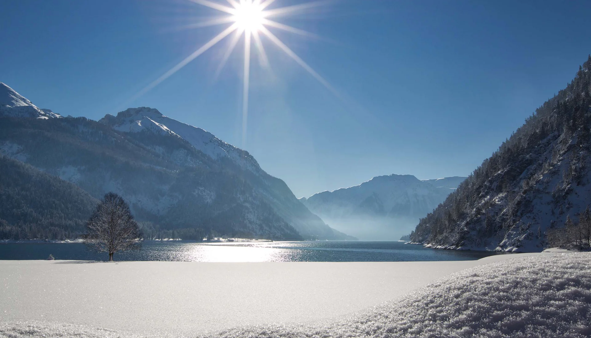 Stammgast werden und Buchau-Taler sammeln Schneebedeckter Bergsee bei Sonnenschein mit Bergen und blauem Himmel