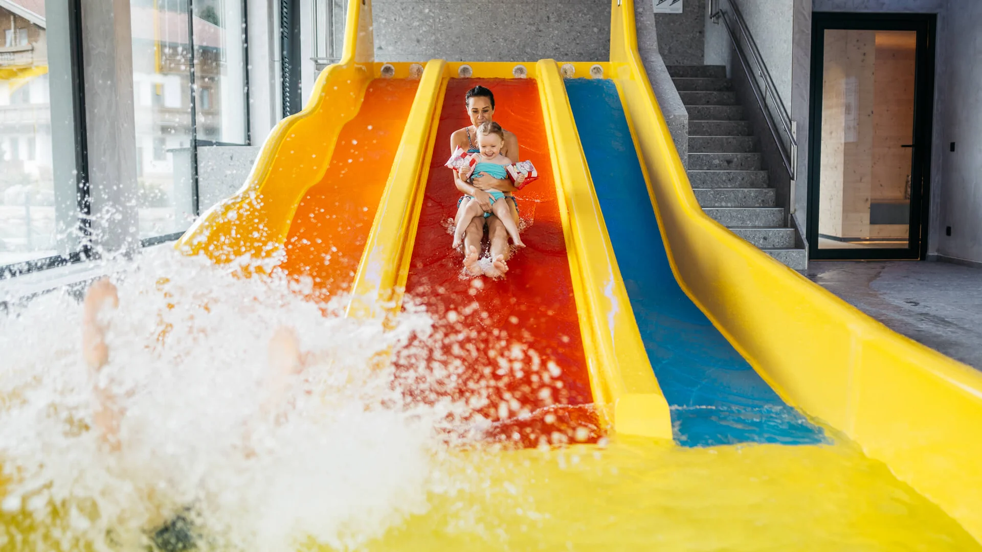 Wasserspaß im Familienhotel in Tirol mit Pool Frau und Kind rutschen zusammen eine gelb-orange Wasserrutsche hinunter im Schwimmbad