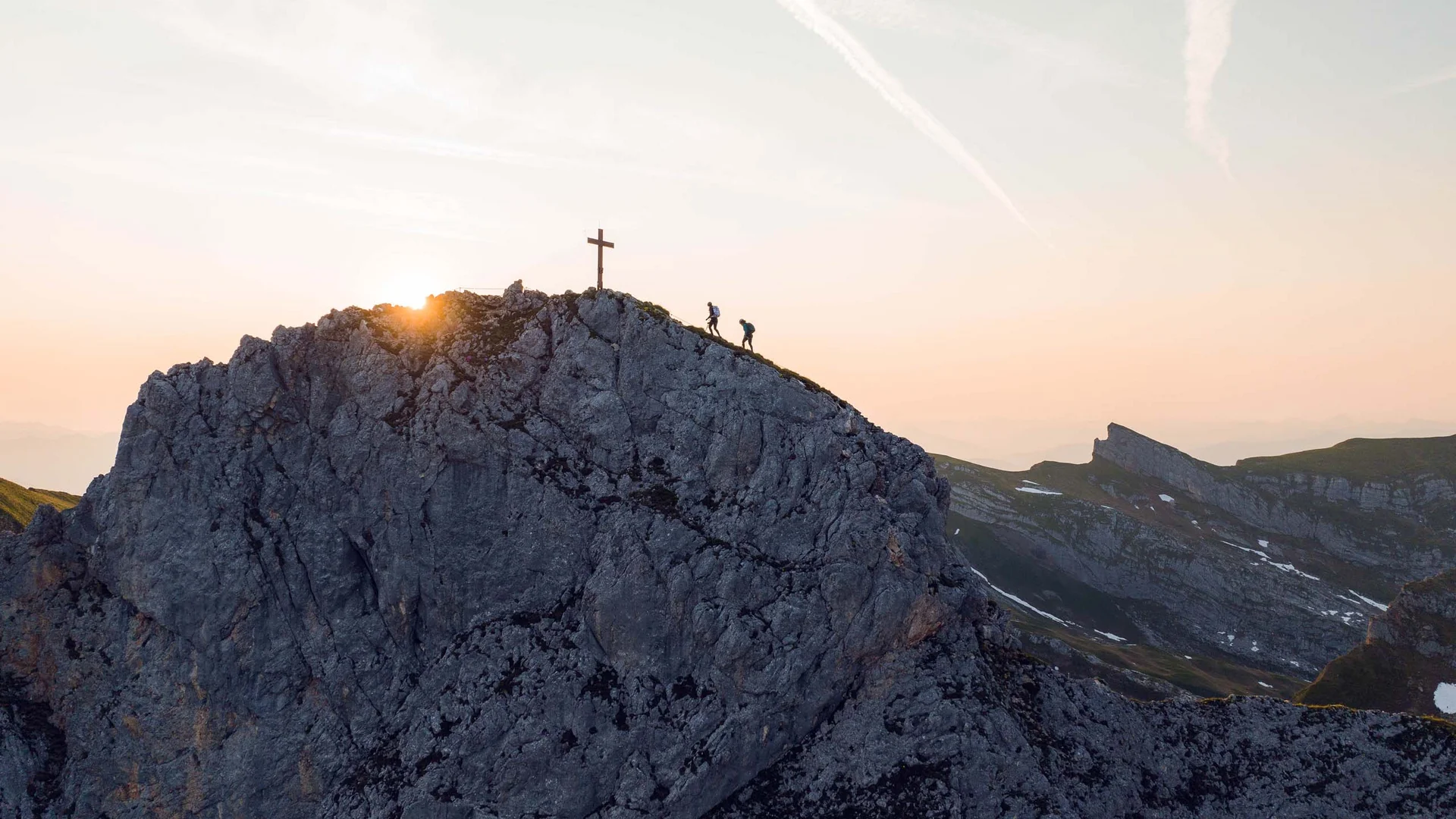 Vom Wanderhotel in Tirol direkt in die Natur Zwei Wanderer steigen bei Sonnenuntergang auf einen Felsen mit Gipfelkreuz