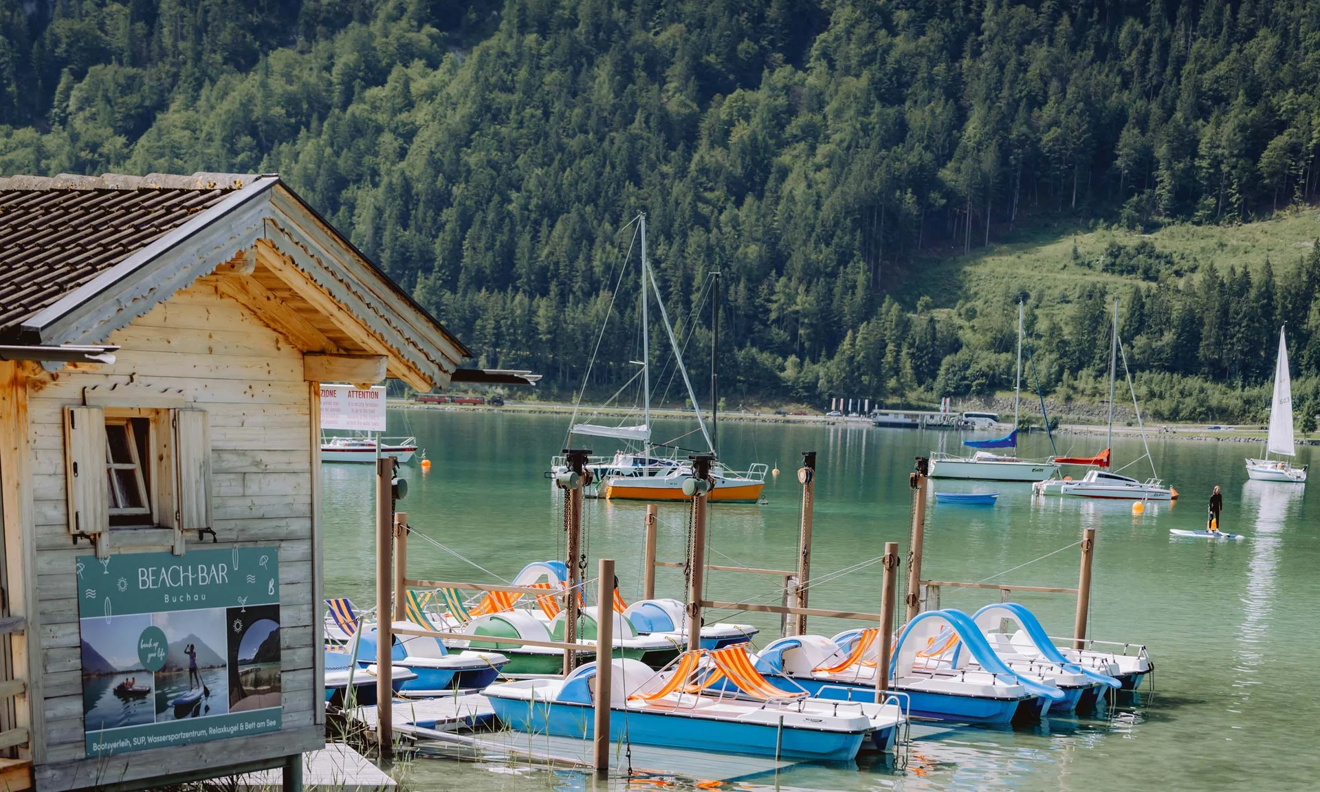 Wassersport auf dem See in Österreich Ruderboote und Segelboote an einem Steg vor bewaldeten Bergen an einem See