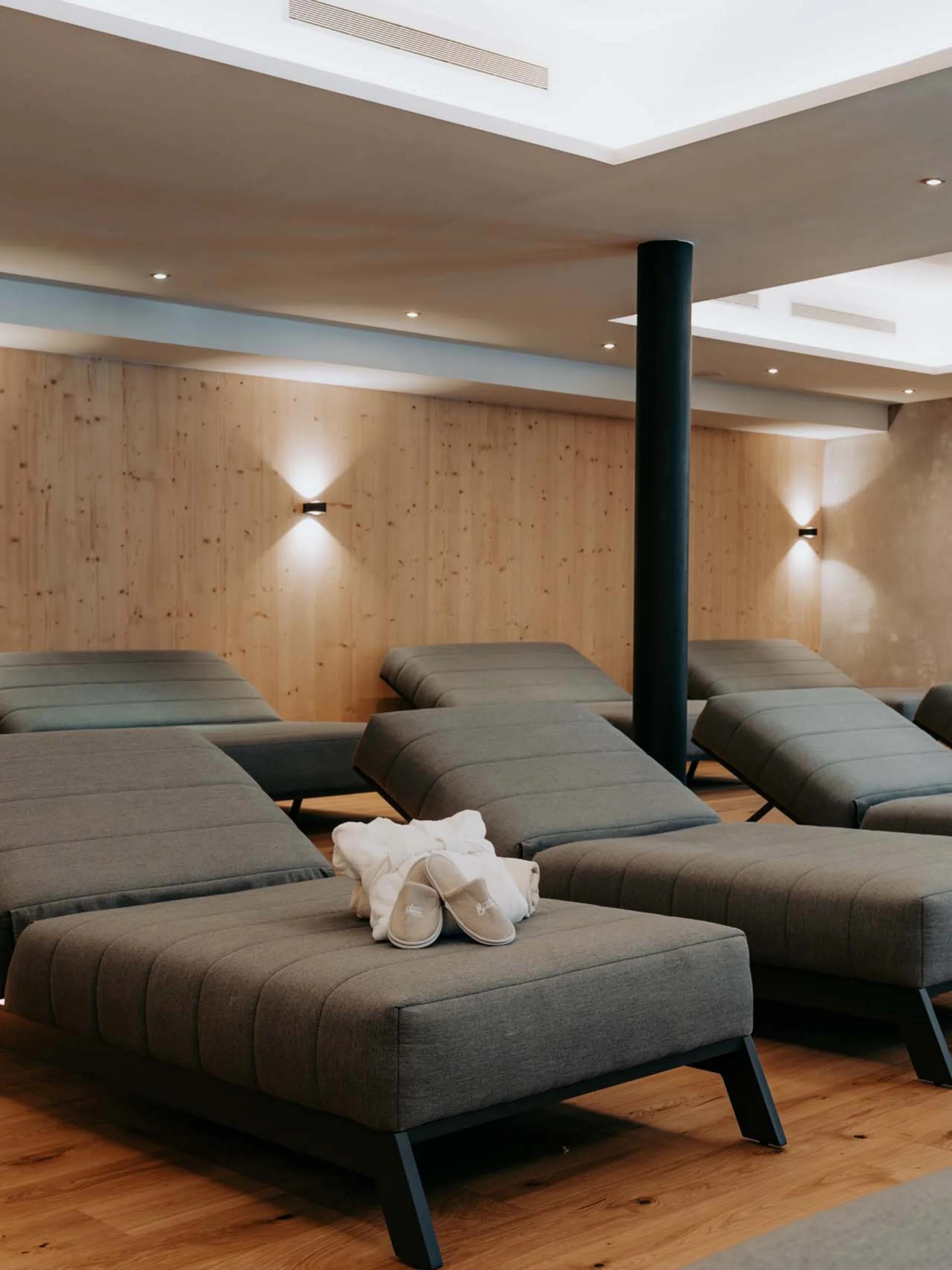 Wir lassen Bilder sprechen. Entspannungsraum mit grauen Liegestühlen, hölzernem Boden und Wandbeleuchtung.