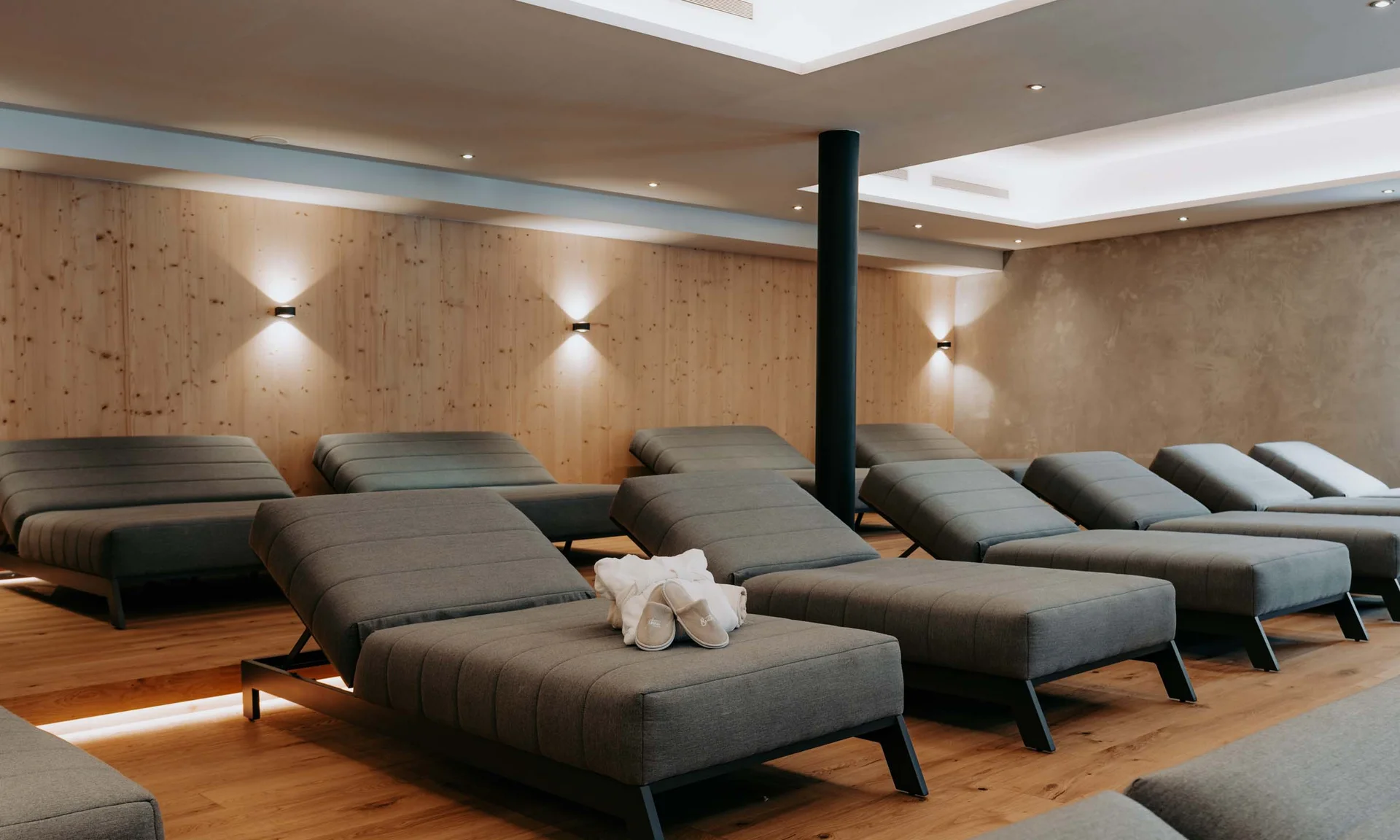 Wir lassen Bilder sprechen. Entspannungsraum mit grauen Liegestühlen, hölzernem Boden und Wandbeleuchtung.