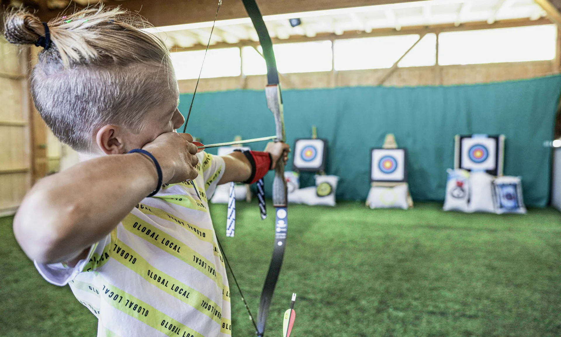 Urlaub im Hotel mit Indoorspielplatz Kind beim Bogenschießen in einer Halle mit Zielscheiben im Hintergrund