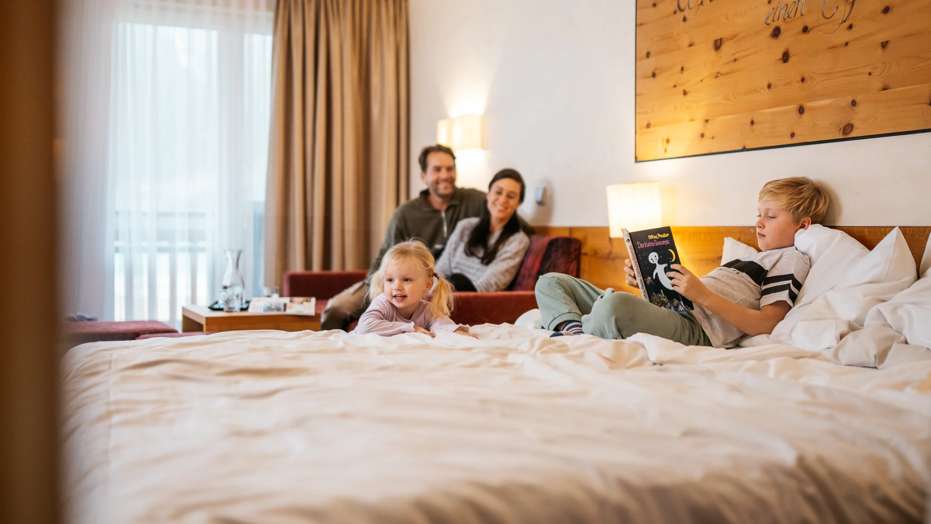 Welcome to our family hotel at Lake Achensee. Family relaxing in hotel room, child reading a book on the bed
