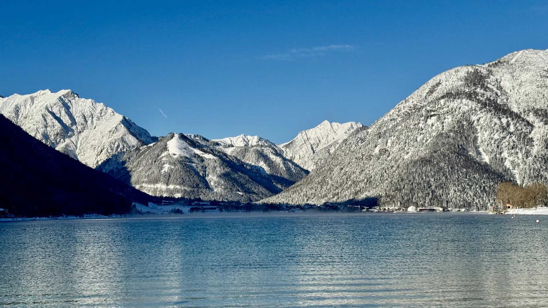Willkommen im Familienhotel Buchau am Achensee Schneebedeckte Berge spiegeln sich im ruhigen Bergsee unter blauem Himmel