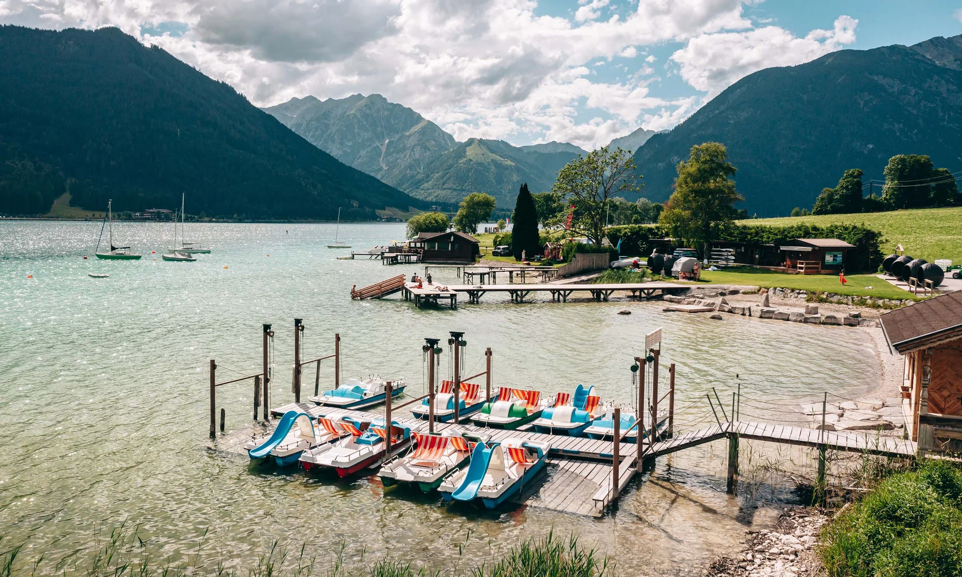 Outdoorspaß im Familienhotel am See in Österreich Tretboote mit Rutschen am Seeufer vor Bergen unter bewölktem Himmel