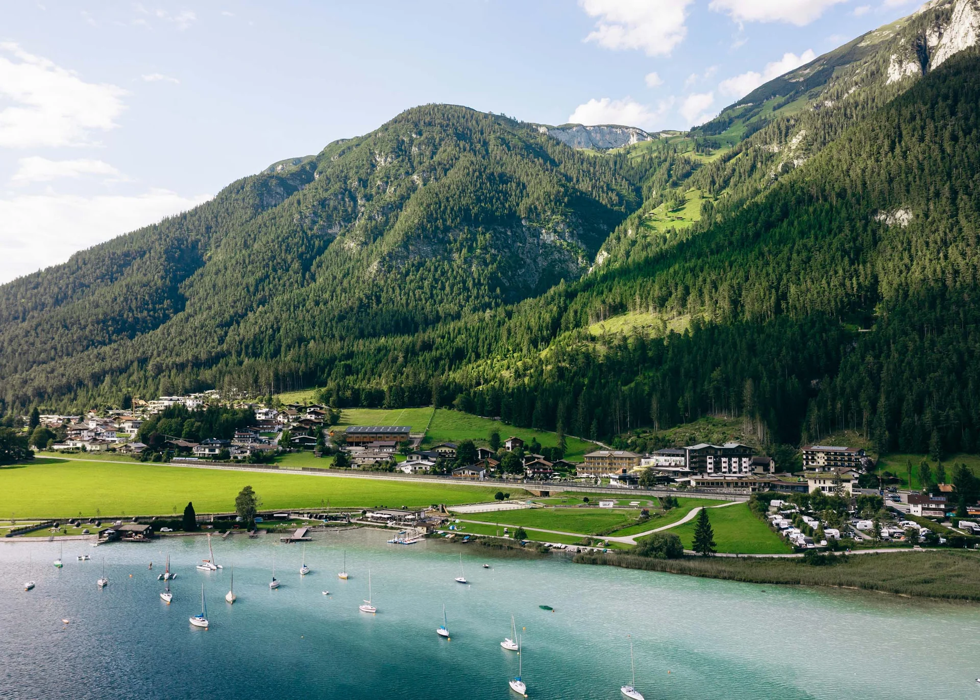 Vom Wanderhotel in Tirol direkt in die Natur Dorf am See mit Segelbooten und bewaldeten Bergen im Hintergrund