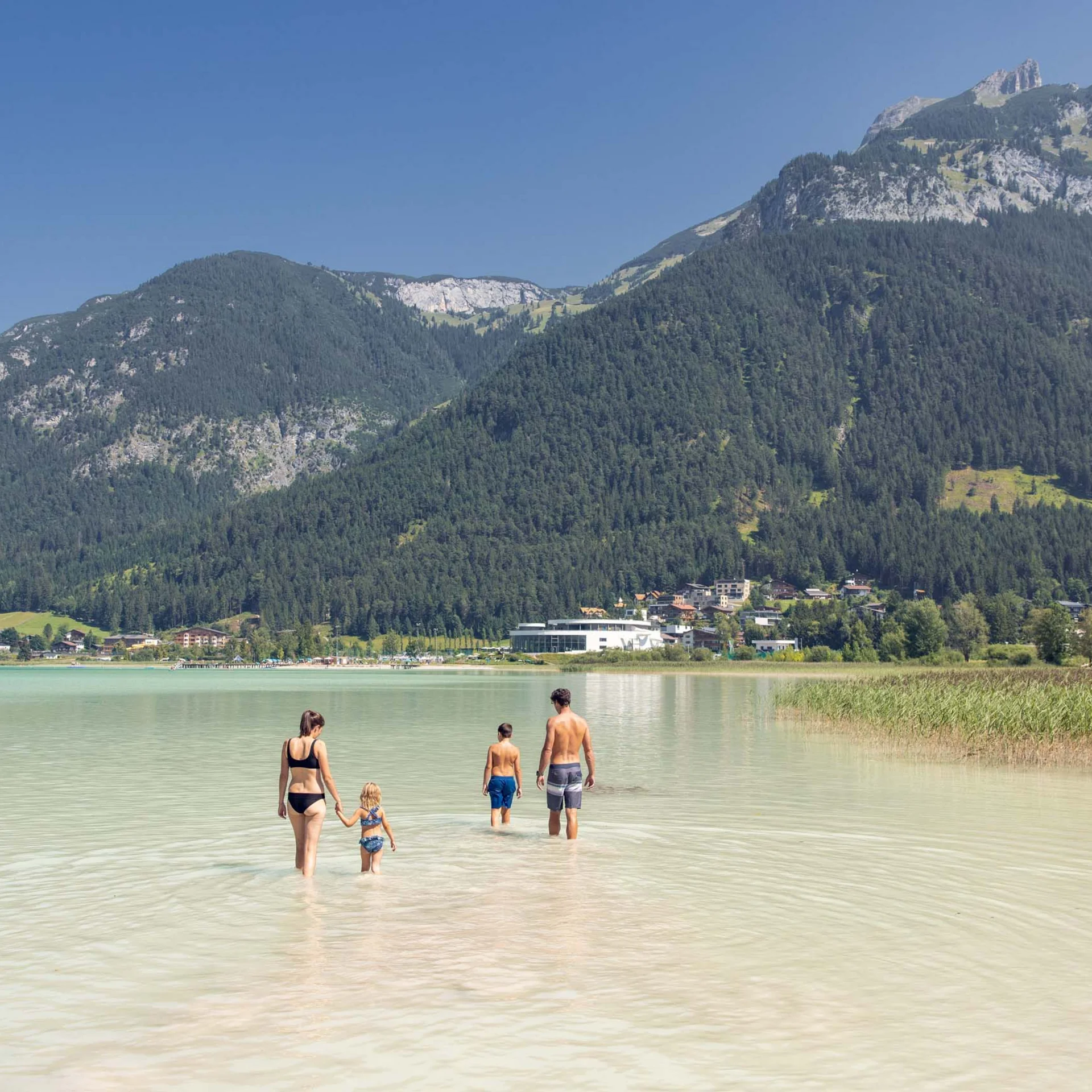 Welcome to our family hotel at Lake Achensee. Family wading in clear mountain lake with mountains and village behind