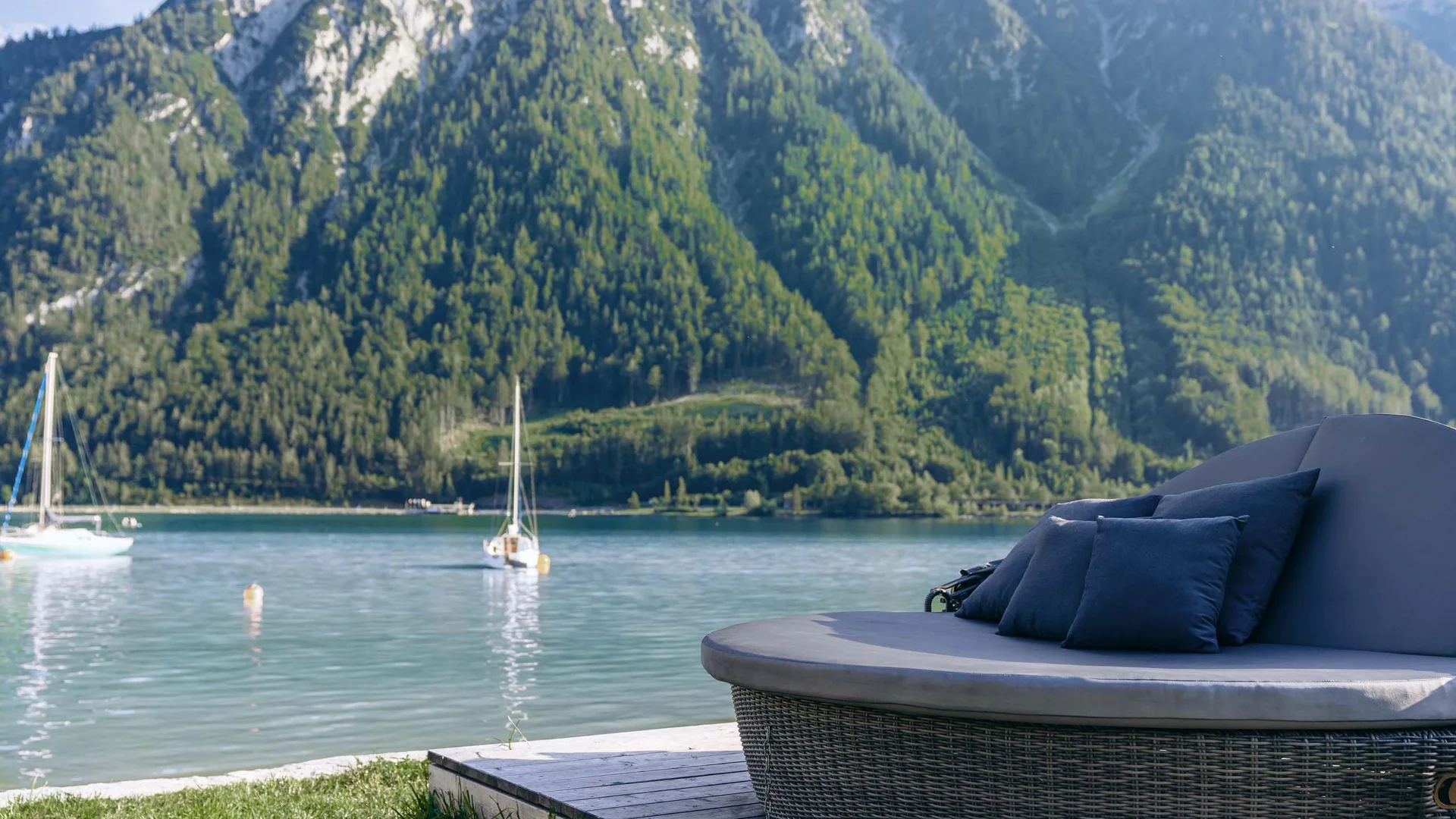 Urlaub im Wellnesshotel mit Kindern in Tirol Rundes Lounge-Sofa mit Kissen am See mit Segelbooten und bewaldeten Bergen im Hintergrund