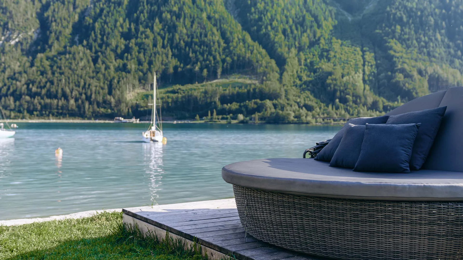 Wasserspaß im Familienhotel in Tirol mit Pool Rundes Lounge-Sofa mit Kissen am See mit Segelbooten und bewaldeten Bergen im Hintergrund