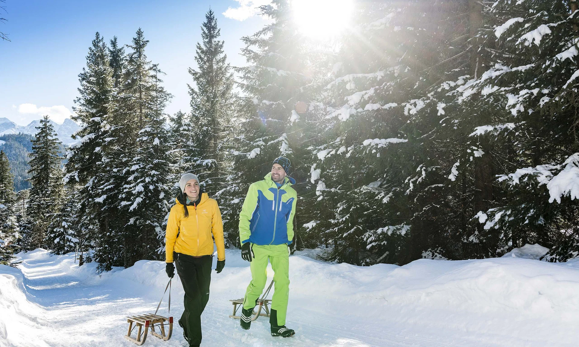 Your unforgettable active lakeside holiday in Austria Couple walking with sleds on snowy forest path in sunlight