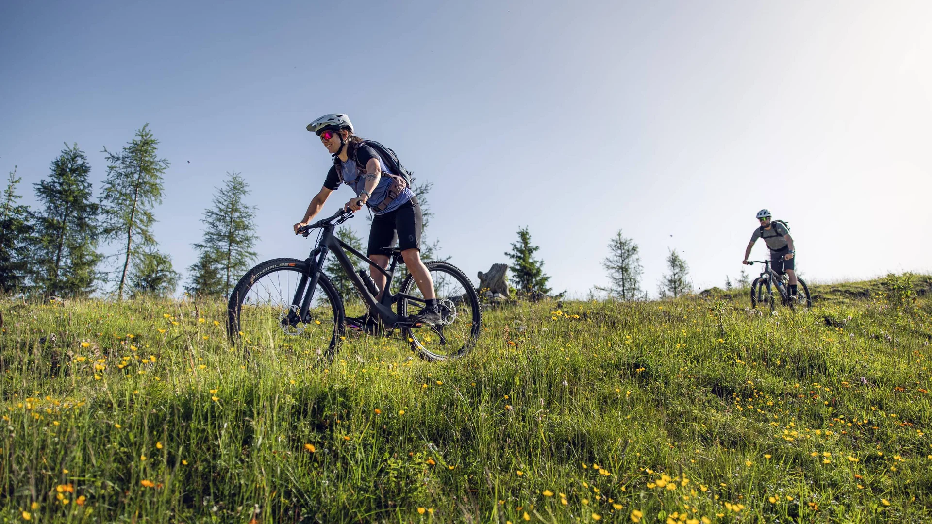 Your unforgettable active lakeside holiday in Austria Two women mountain biking on a flower-covered hill on a sunny day