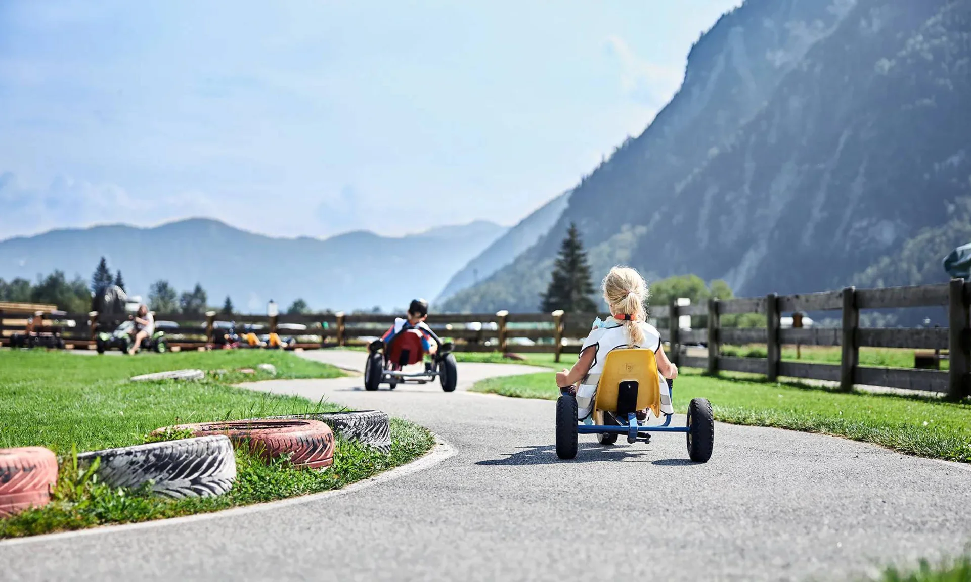 Welcome to our family hotel at Lake Achensee. Children riding pedal cars on a winding track in a rural mountain setting
