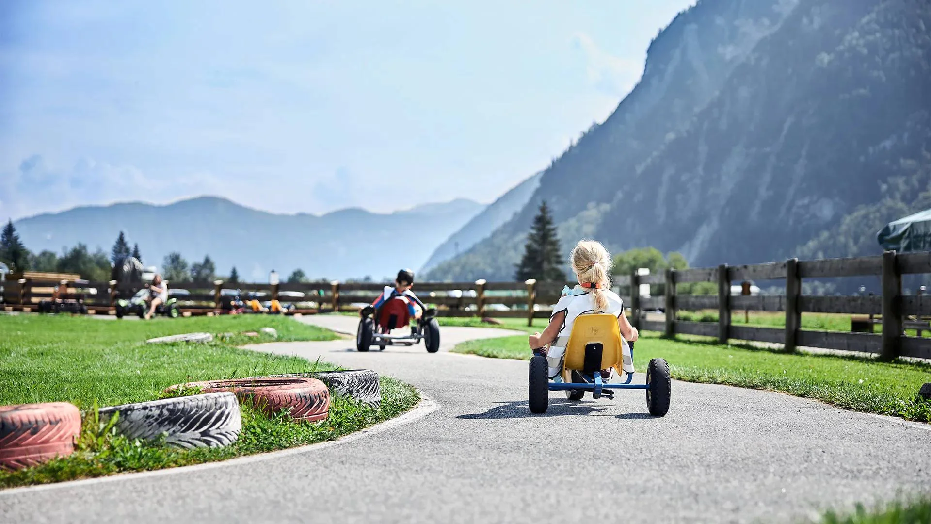 Outdoorspaß im Familienhotel am See in Österreich Kinder fahren Tretautos auf einer kurvigen Bahn in ländlicher Berglandschaft