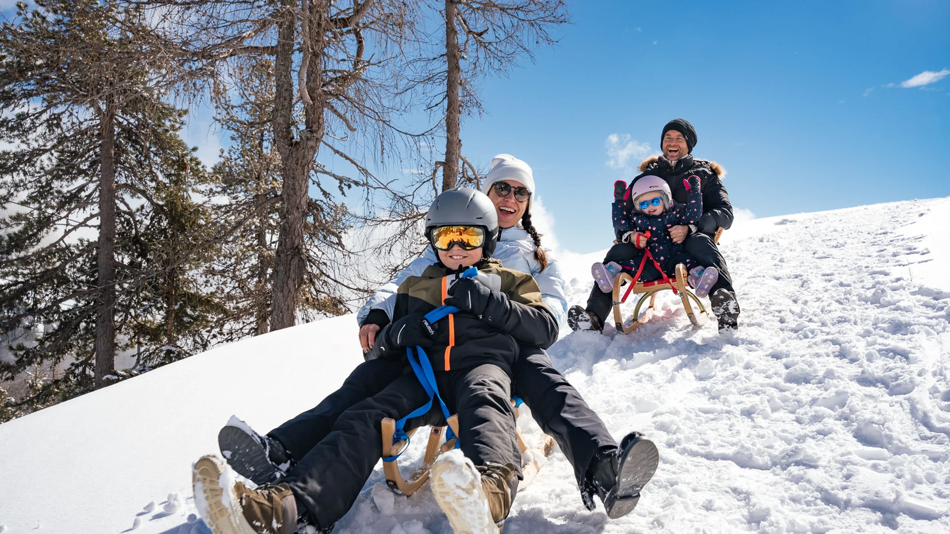 Willkommen im Familienhotel Buchau am Achensee Familienmitglieder rodeln lachend den verschneiten Hügel bei sonnigem Wetter hinunter