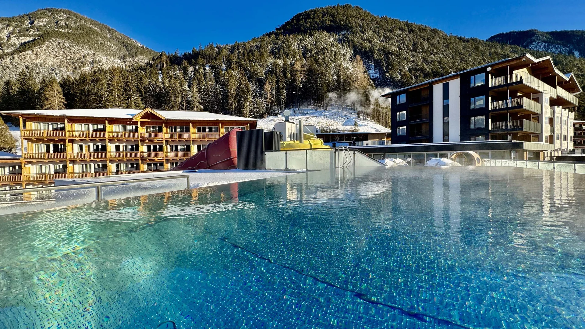 Willkommen im Familienhotel Buchau am Achensee Beheizter Außenpool mit Bergkulisse und Gebäuden im Hintergrund unter klarem Himmel