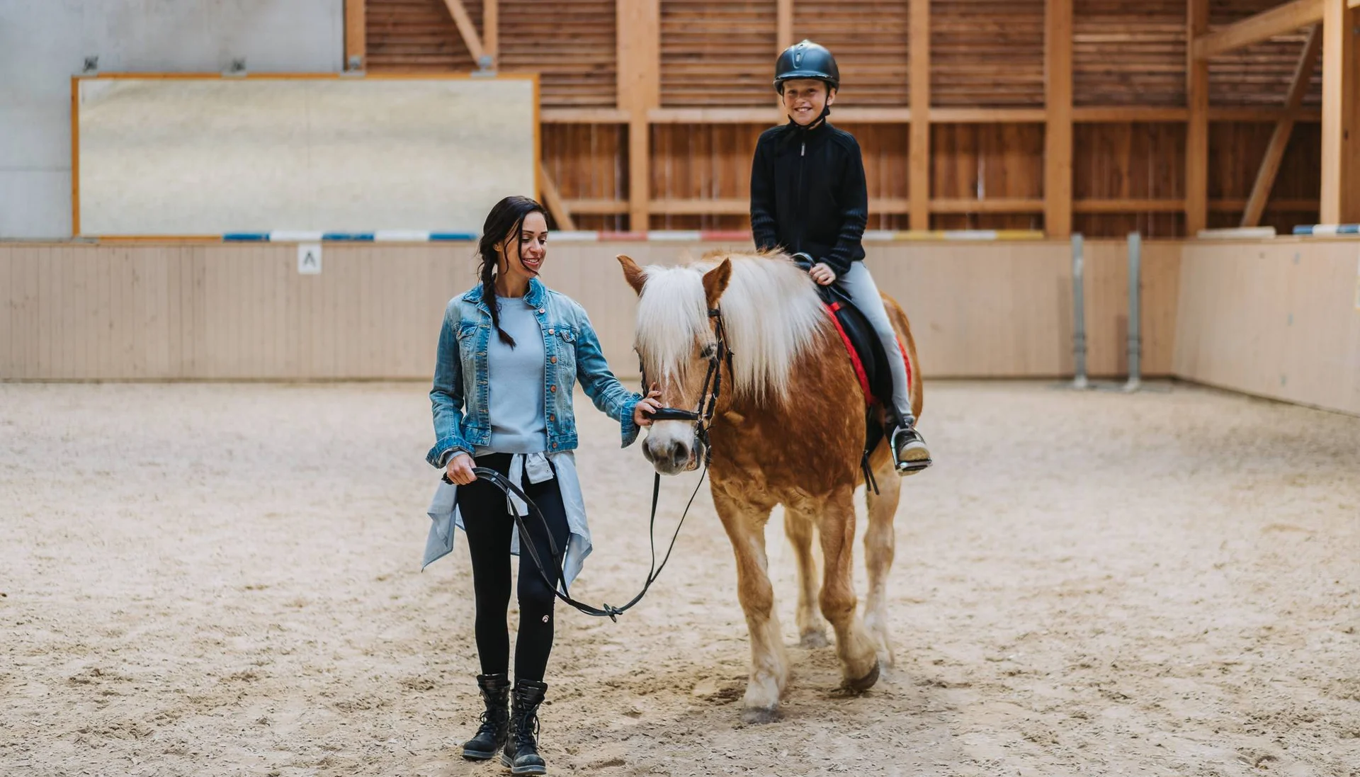 Urlaub im Hotel mit Indoorspielplatz Frau führt Pony, Kind reitet mit Helm in Reithalle