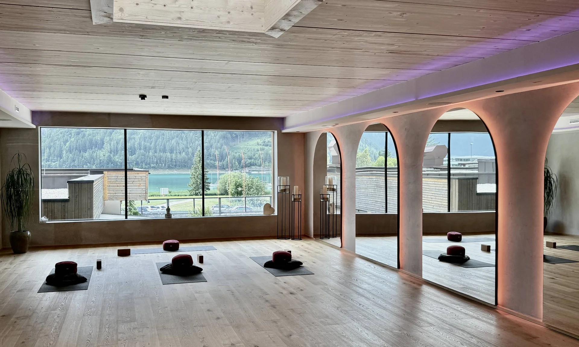 Wir lassen Bilder sprechen. Helles Yoga-Studio mit Holzboden, Meditationskissen und großer Fensterfront mit Seeblick