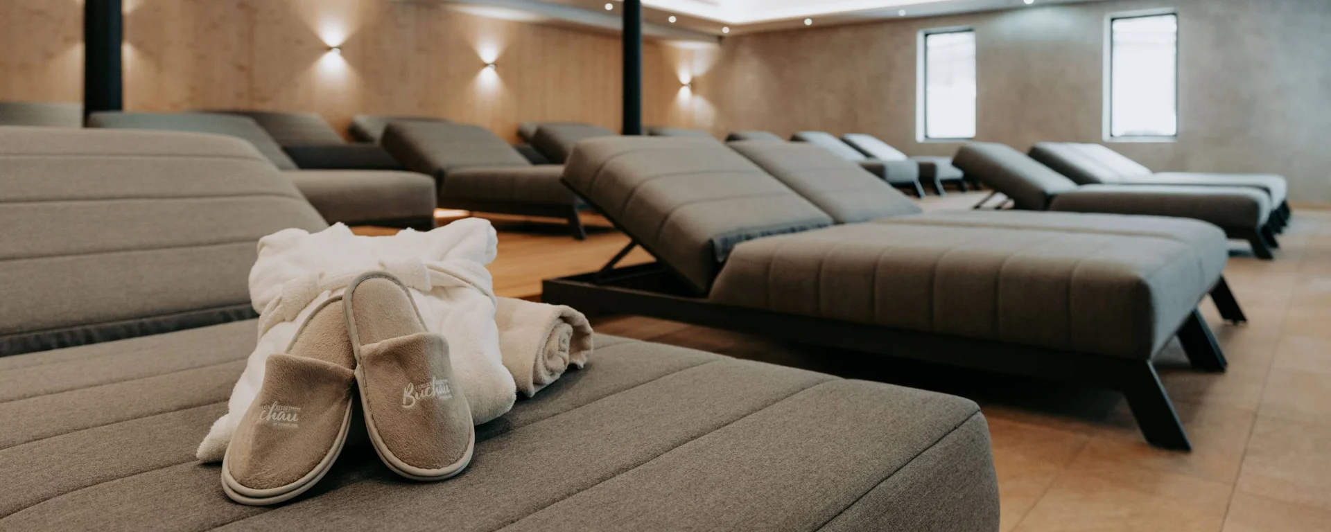 Ab ins Wellnesshotel in Tirol, in den Infinitypool! Entspannungsraum mit grauen Liegen, Bademantel, Handtuch und Hausschuhen