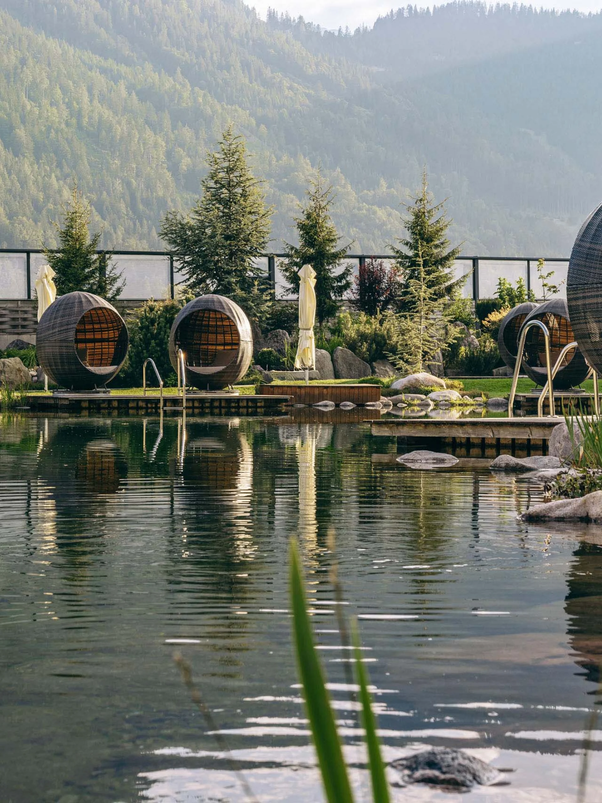 Wir lassen Bilder sprechen. Natürlicher Pool mit runden Sitzpods in einer Gartenanlage mit Bergen im Hintergrund